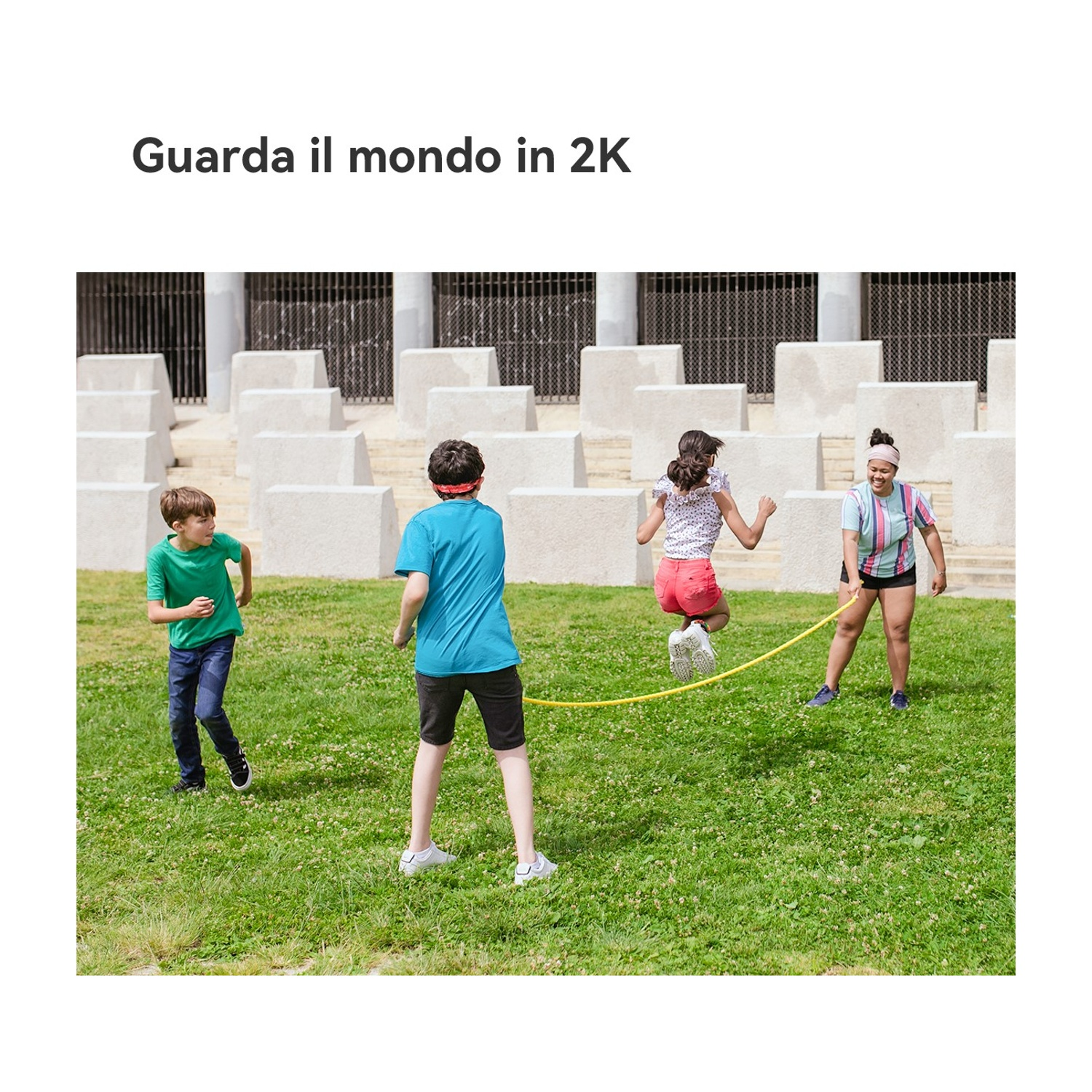 Quattro bambini giocano a saltare la corda sull'erba. Uno salta, due girano la corda e uno guarda. Lo sfondo presenta blocchi di cemento.