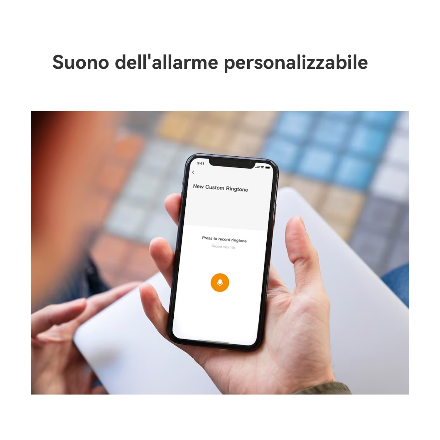 Una persona tiene in mano un telefono che mostra 'New Custom Ringtone' con un pulsante di registrazione. Il telefono è appoggiato su un laptop.