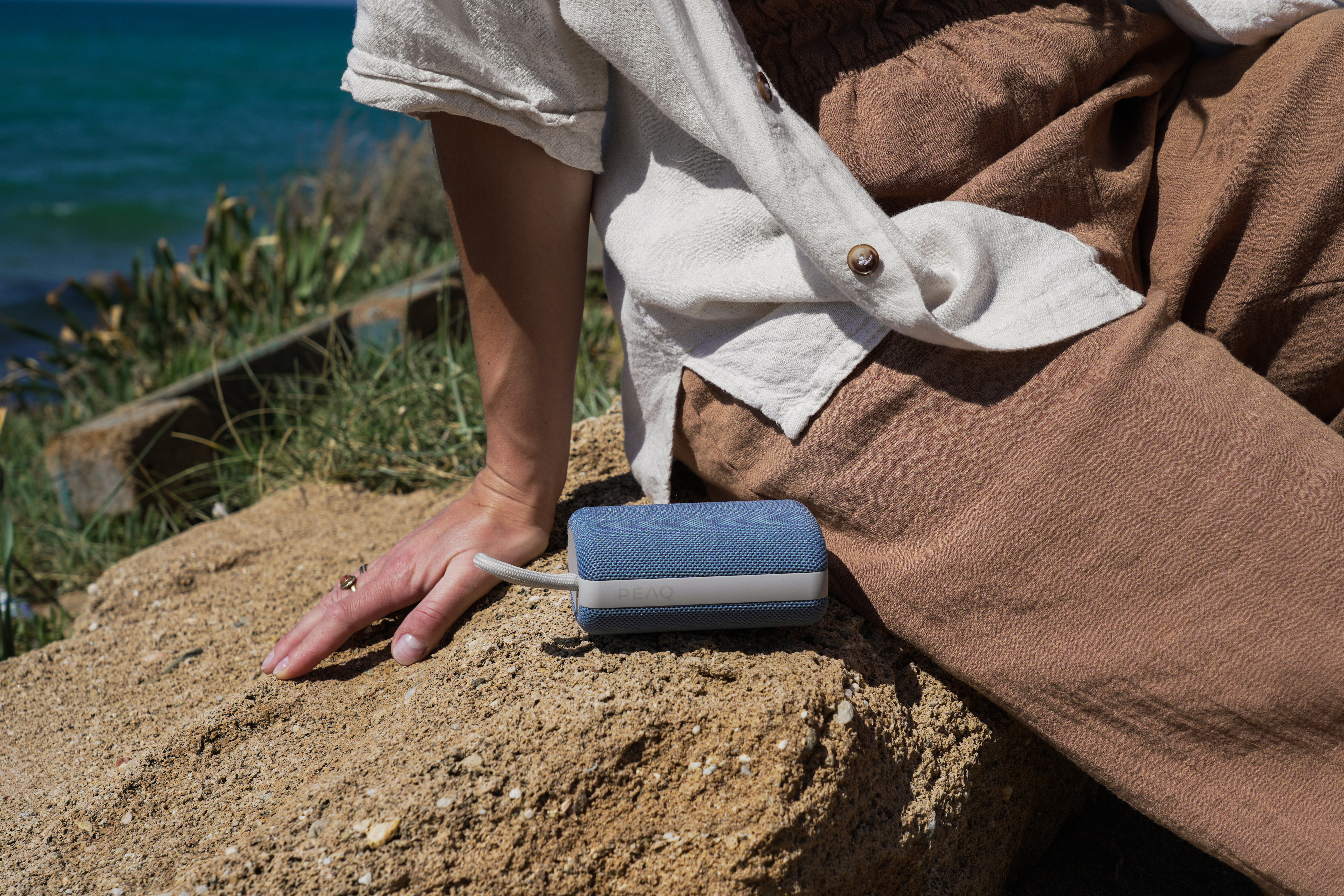Une personne pose sa main près d'un haut-parleur bleu sur du sable. La mer et la verdure sont en arrière-plan.