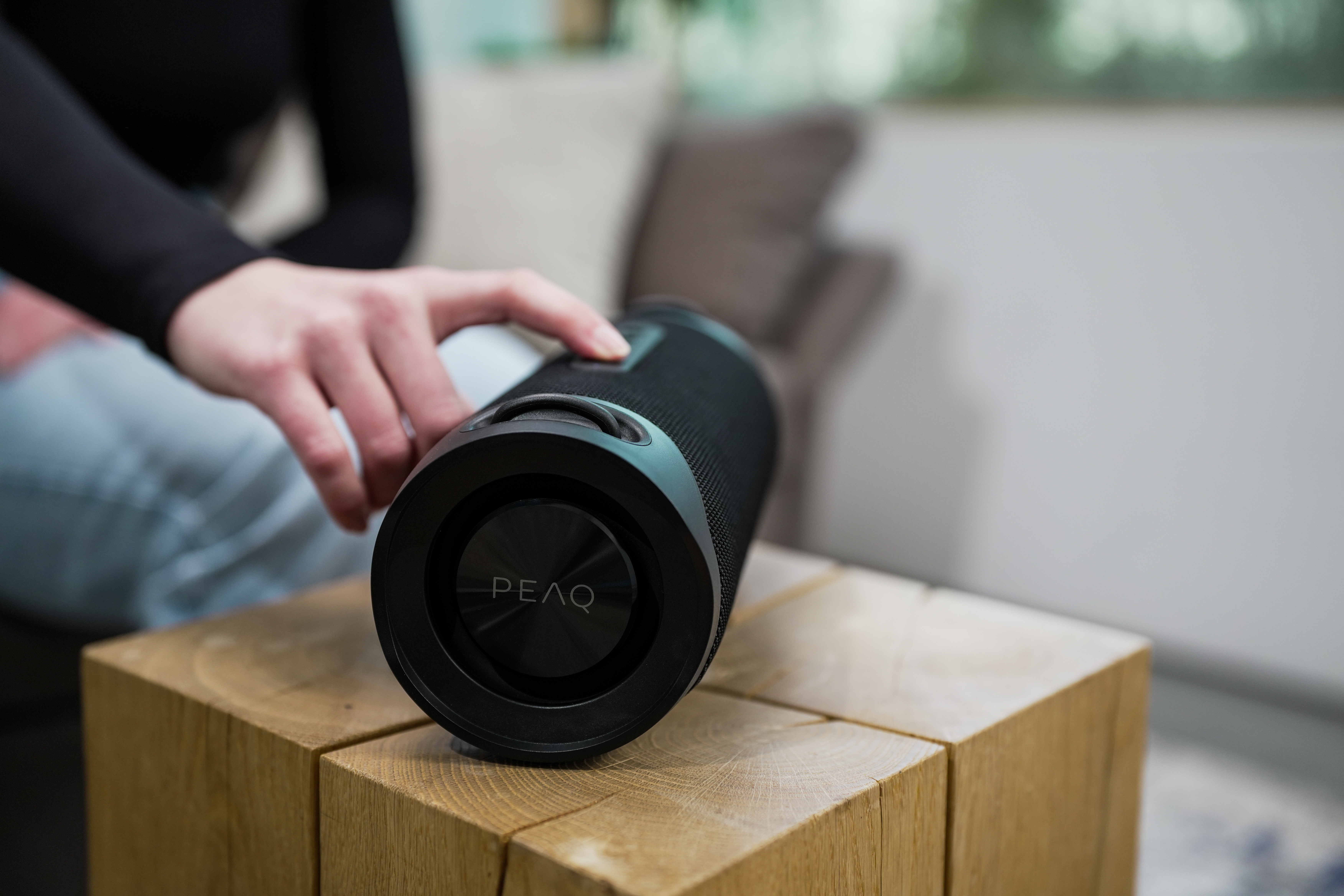 Une main touche un haut-parleur cylindrique noir sur un bloc de bois. Le haut-parleur porte la marque PEAQ.