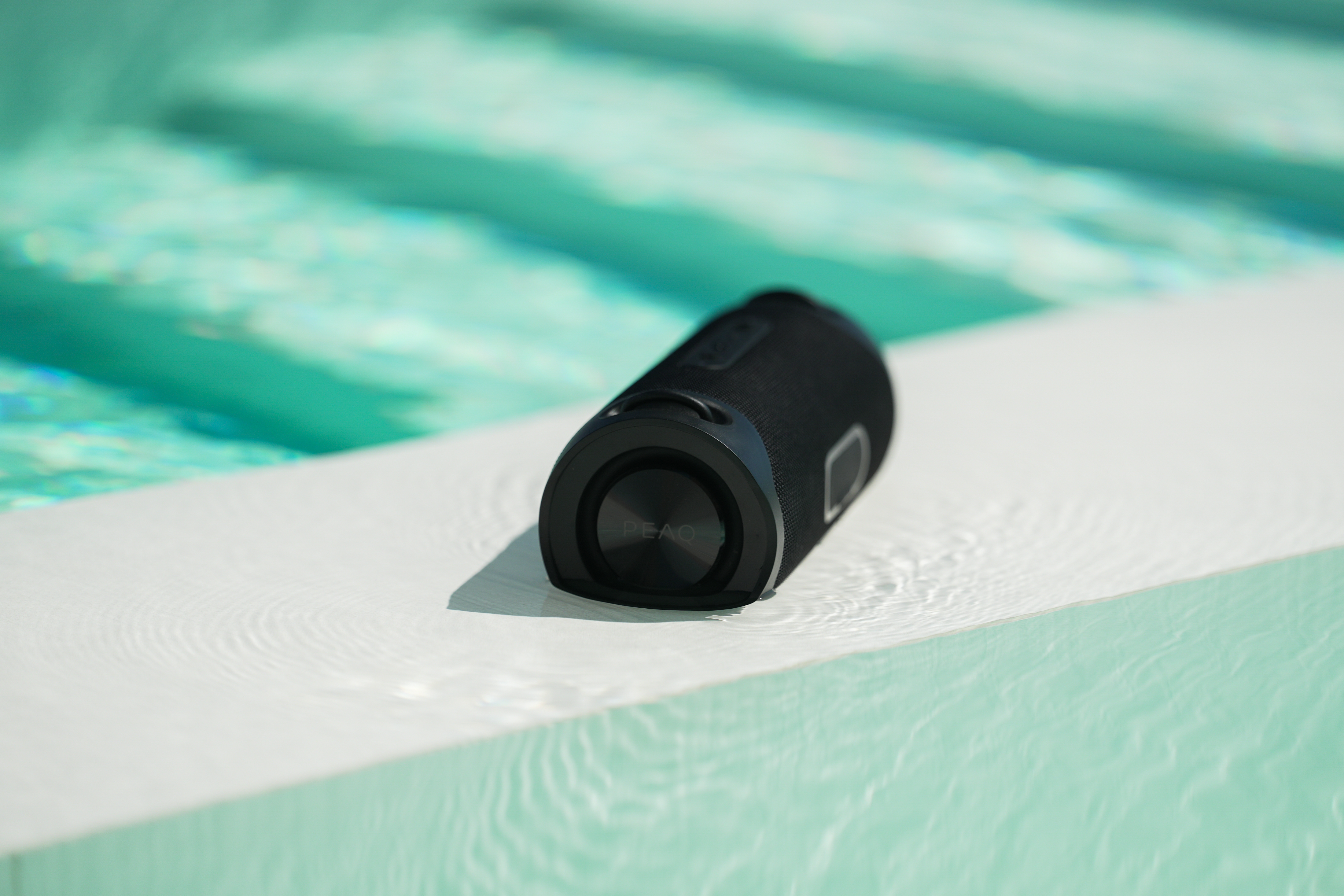 Un haut-parleur noir sur une surface blanche, avec de l'eau autour. L'arrière-plan est une piscine.