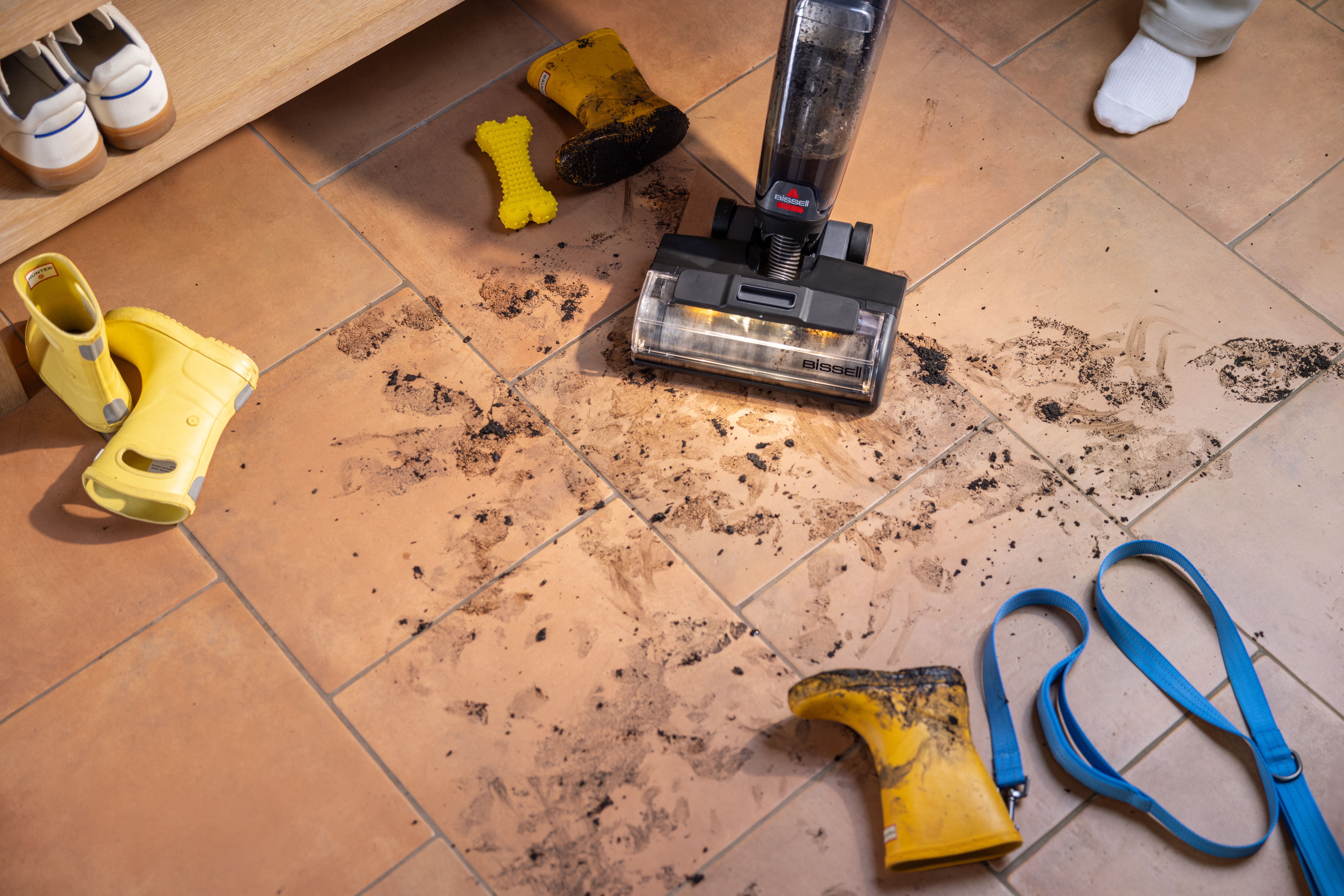 Podłoga z brudem, żółte buty, odkurzacz i niebieska smycz. Buty po lewej stronie. Widoczna stopa.