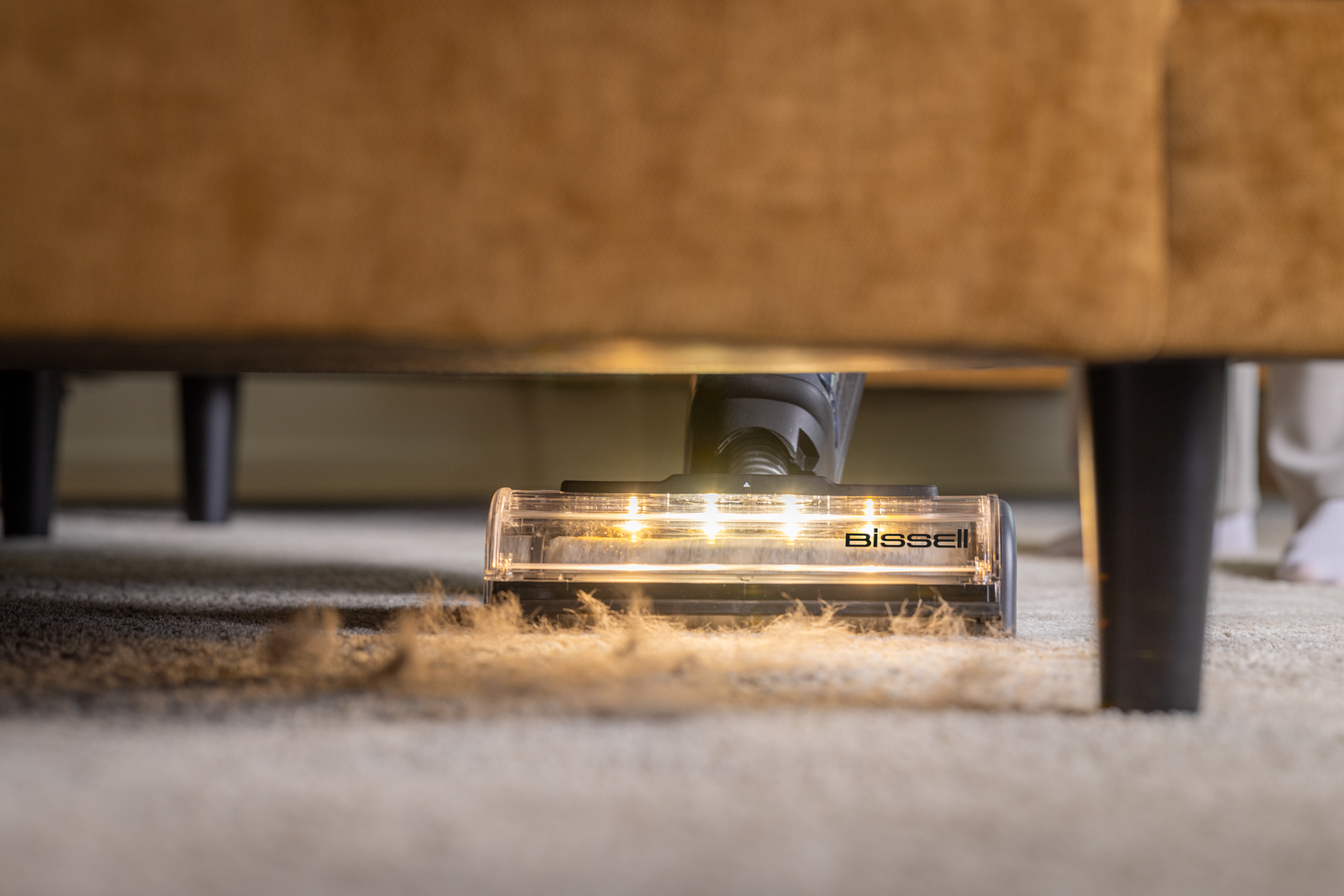 Odkurzacz czyści dywan pod brązową sofą. Widoczne jest logo Bissell.