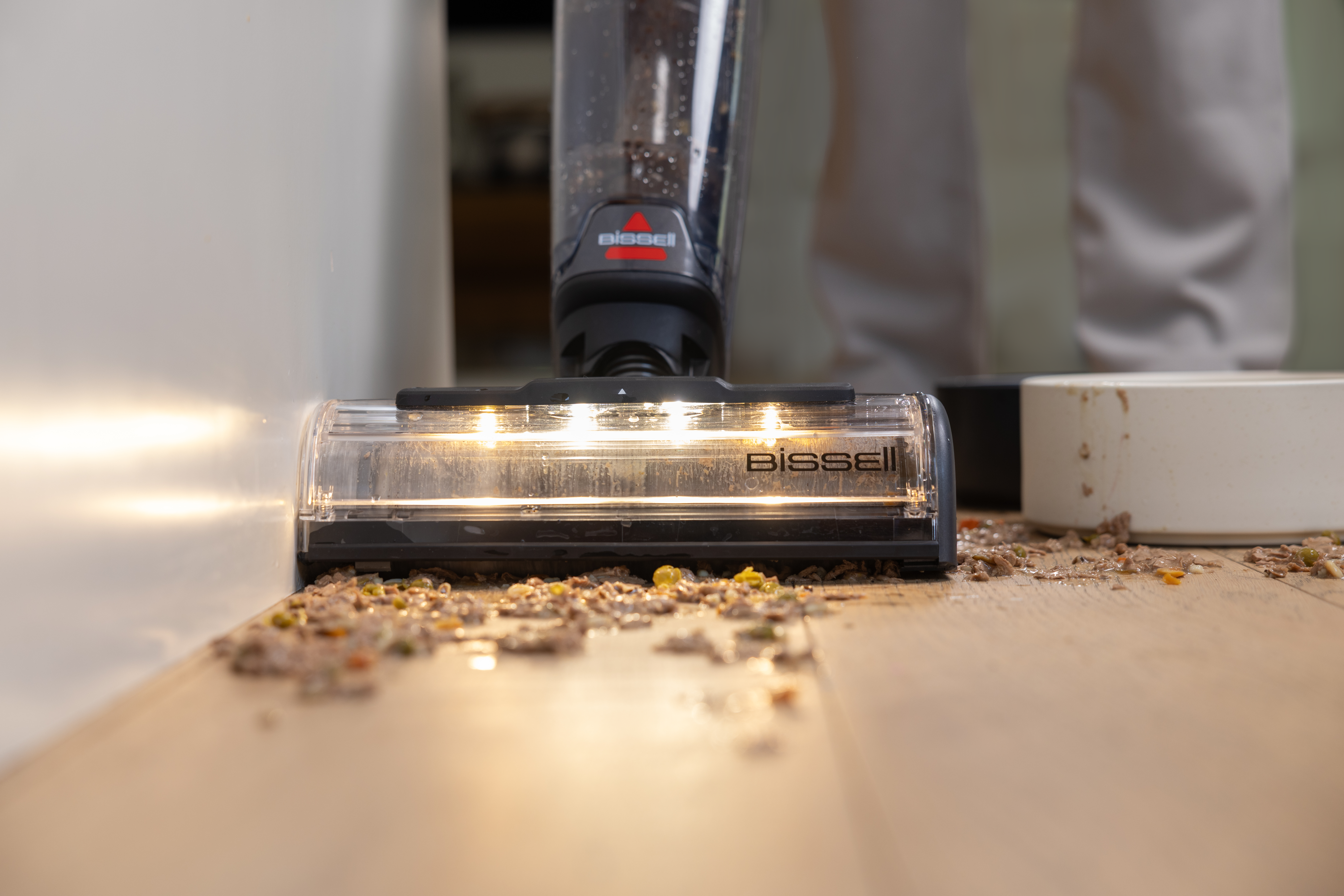 Odkurzacz Bissell sprząta rozlane jedzenie dla zwierząt na drewnianej podłodze. Odkurzacz jest czarny i przezroczysty.