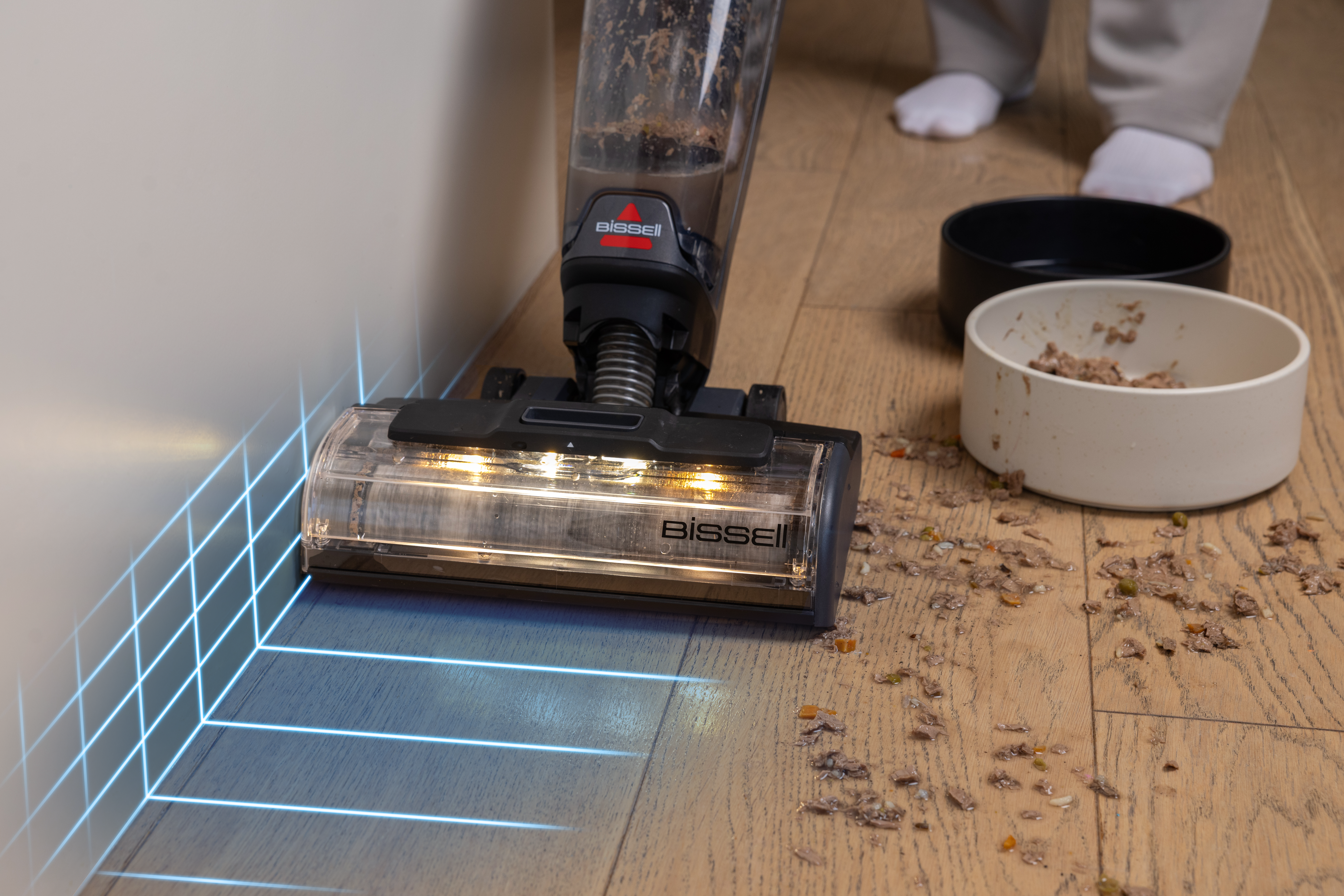 Odkurzacz Bissell czyści drewnianą podłogę z rozrzuconym jedzeniem. Stopy osoby i miski w tle.