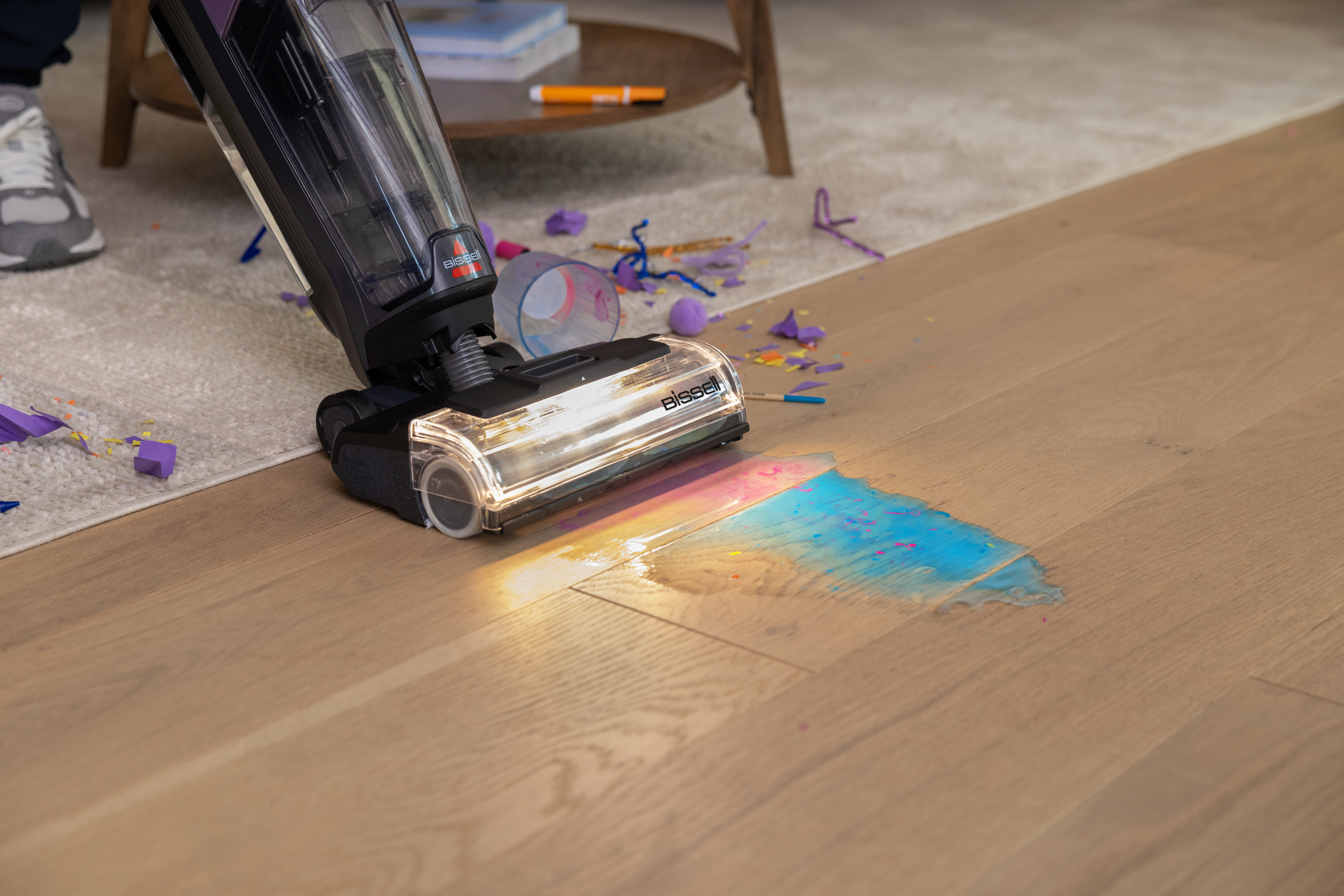 Odkurzacz czyszczący kolorową plamę na drewnianej podłodze, z rozrzuconymi odłamkami.