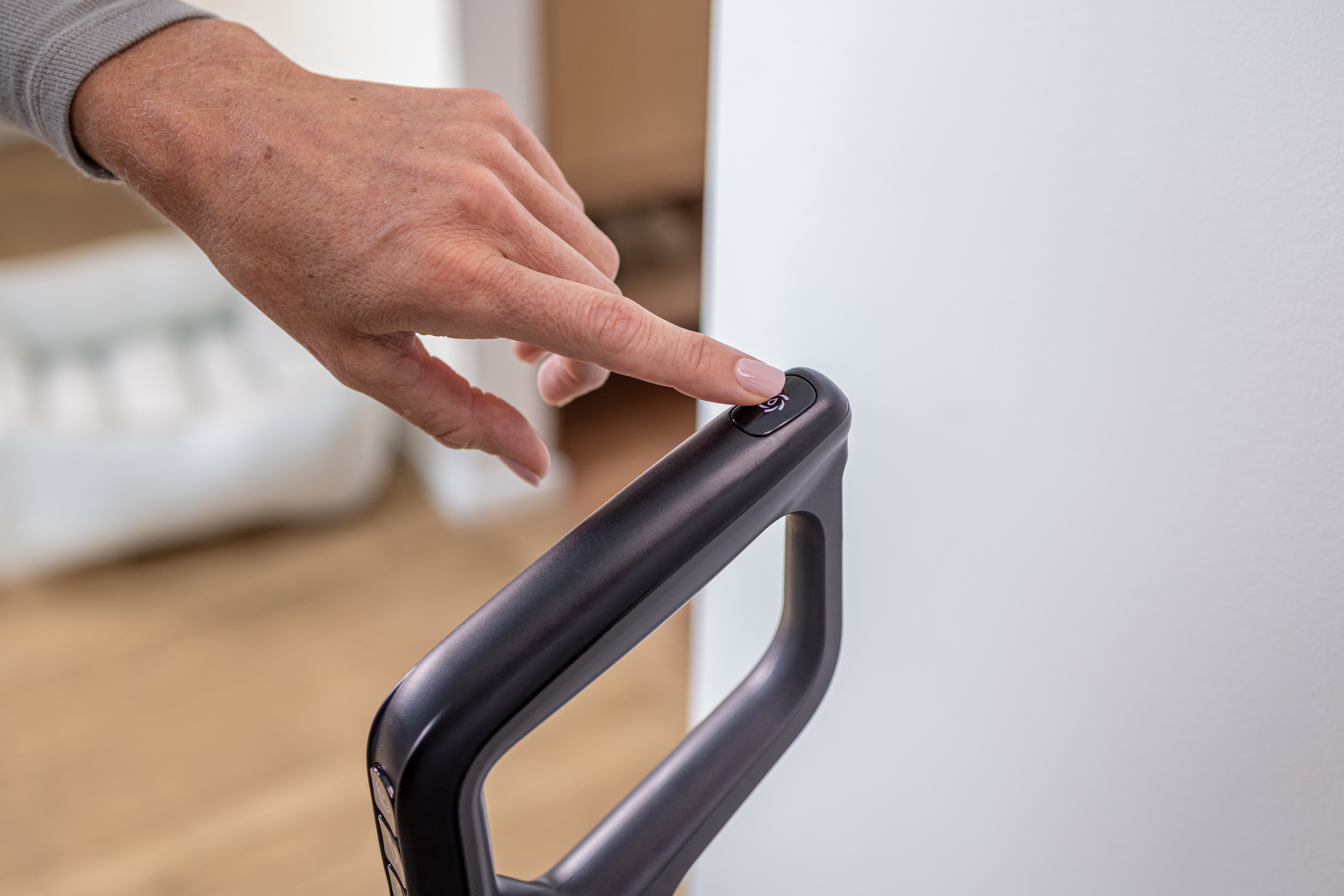 Dłoń naciska przycisk zasilania na czarnym uchwycie odkurzacza, na tle białej ściany.