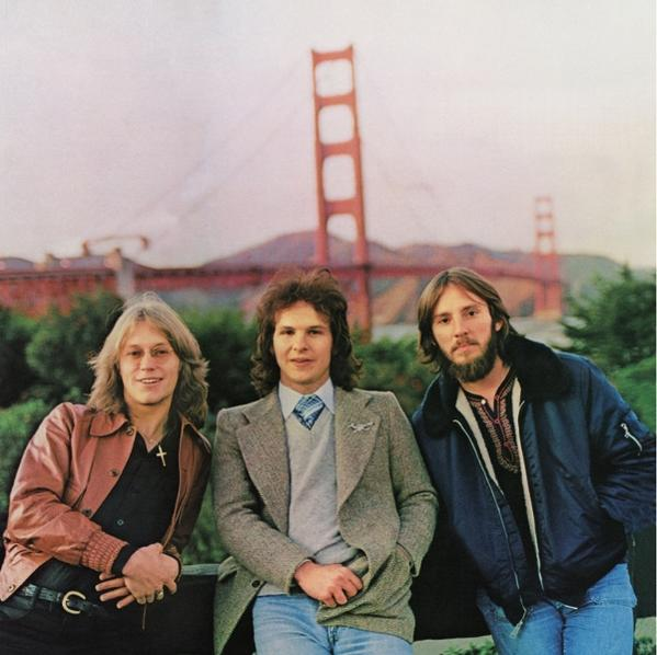 Drei Männer posieren vor der Golden Gate Bridge. Sie tragen Mäntel und Jacken, die Brücke ist rot.