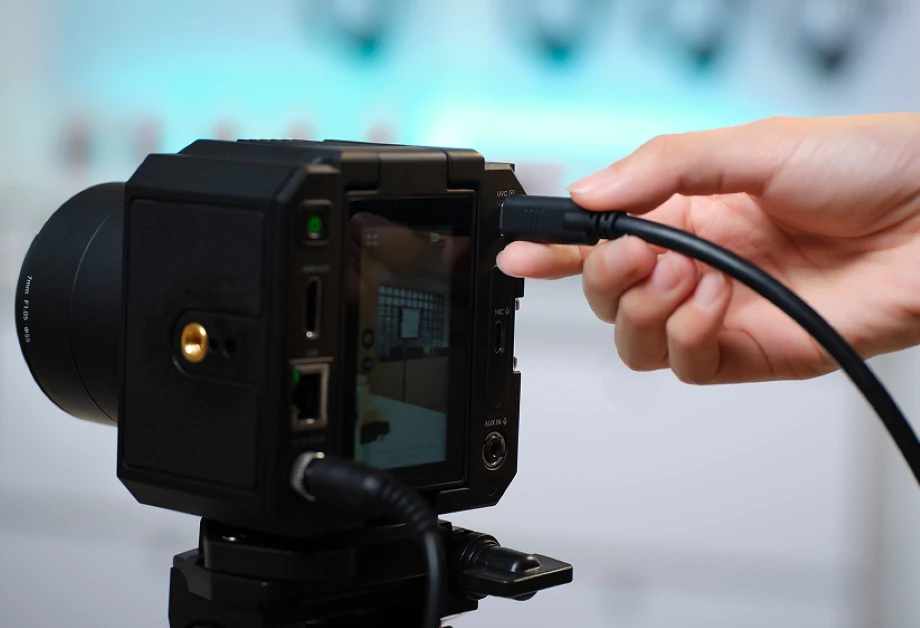Osoba podłącza czarny kabel do czarnego aparatu. Aparat jest na statywie. Tło jest rozmyte.