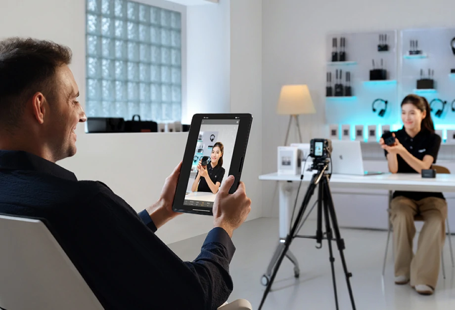 Mężczyzna trzyma tablet z kobietą z aparatem. Kobieta siedzi przy biurku, widoczny jest sprzęt.