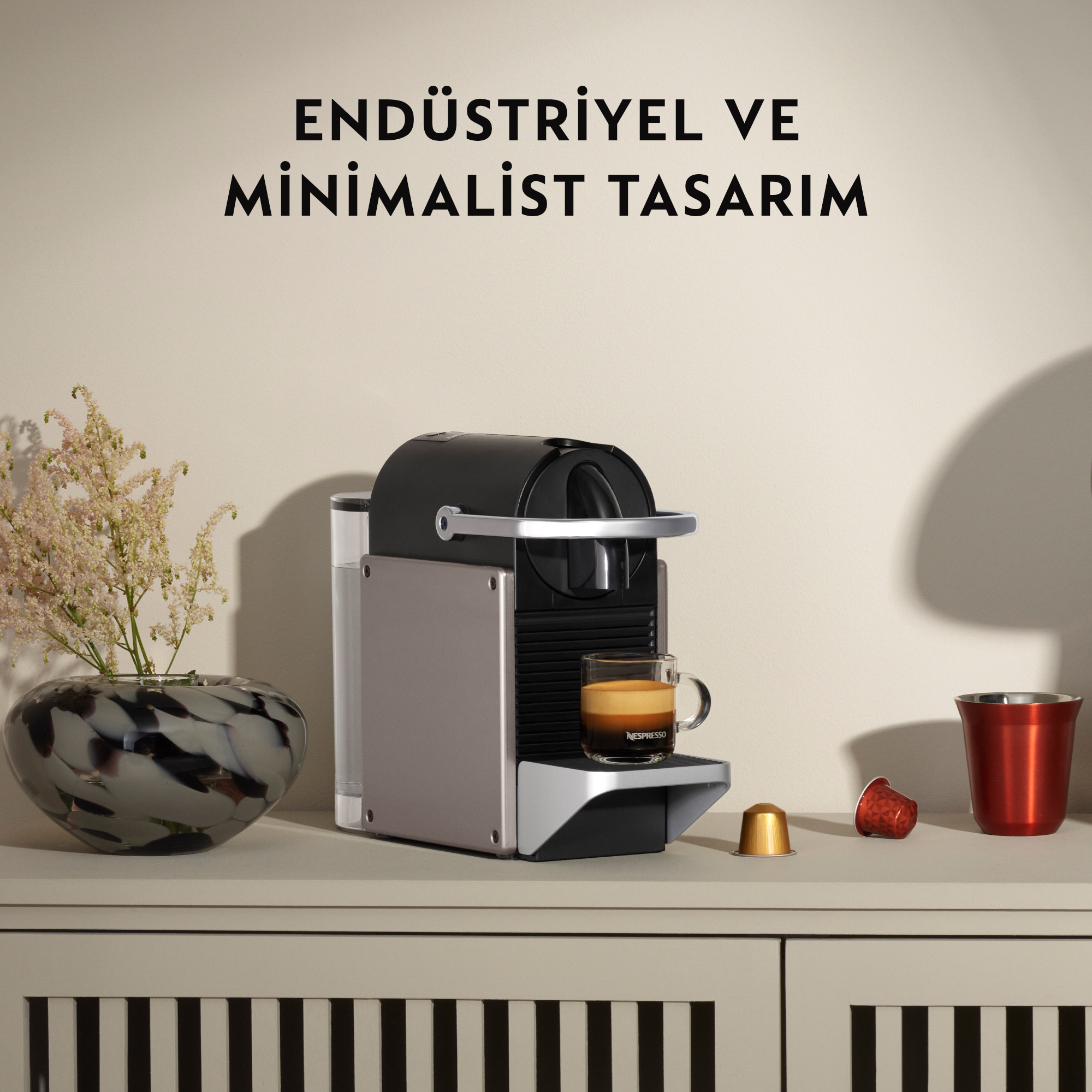 Bir fincan kahve ile bir Nespresso makinesi. Solda çiçekli bir vazo. Sağda kırmızı ve altın kapsüller.