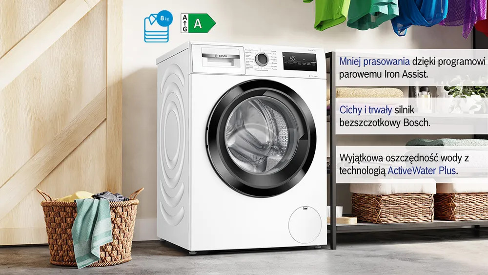 Biała pralka w pokoju. Kosz z ubraniami na pierwszym planie. Półki z ręcznikami po prawej stronie.