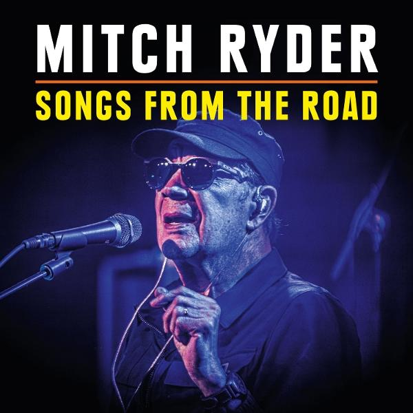 Mitch Ryder singt in ein Mikrofon. Er trägt eine Sonnenbrille, eine schwarze Mütze und ein dunkles Hemd. Der Hintergrund ist schwach beleuchtet.
