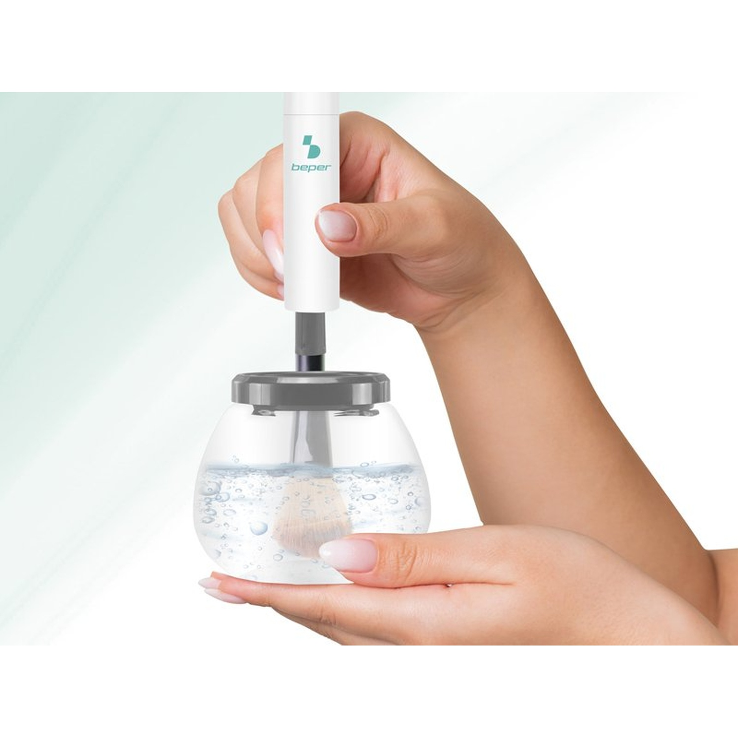 A person holds a makeup brush cleaner. The device has a water-filled glass with a brush inside. The background is white.