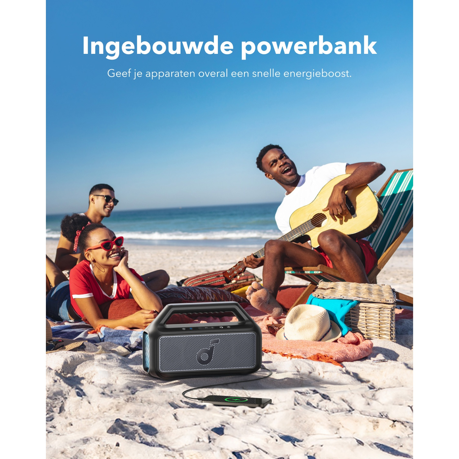 Beach scene: Man plays guitar, friends relax. A portable speaker, hat, and picnic basket are on the sand. Clear blue sky.
