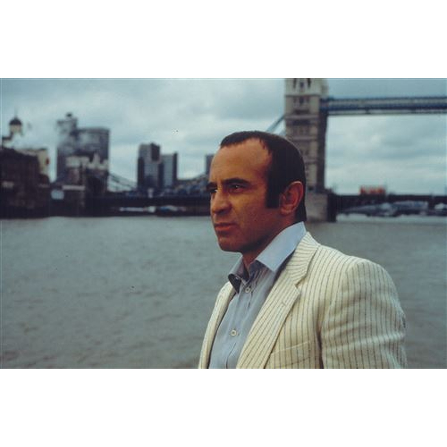 Un homme en veste rayée se tient près d'une rivière, avec le Tower Bridge et des bâtiments de la ville en arrière-plan sous un ciel nuageux.