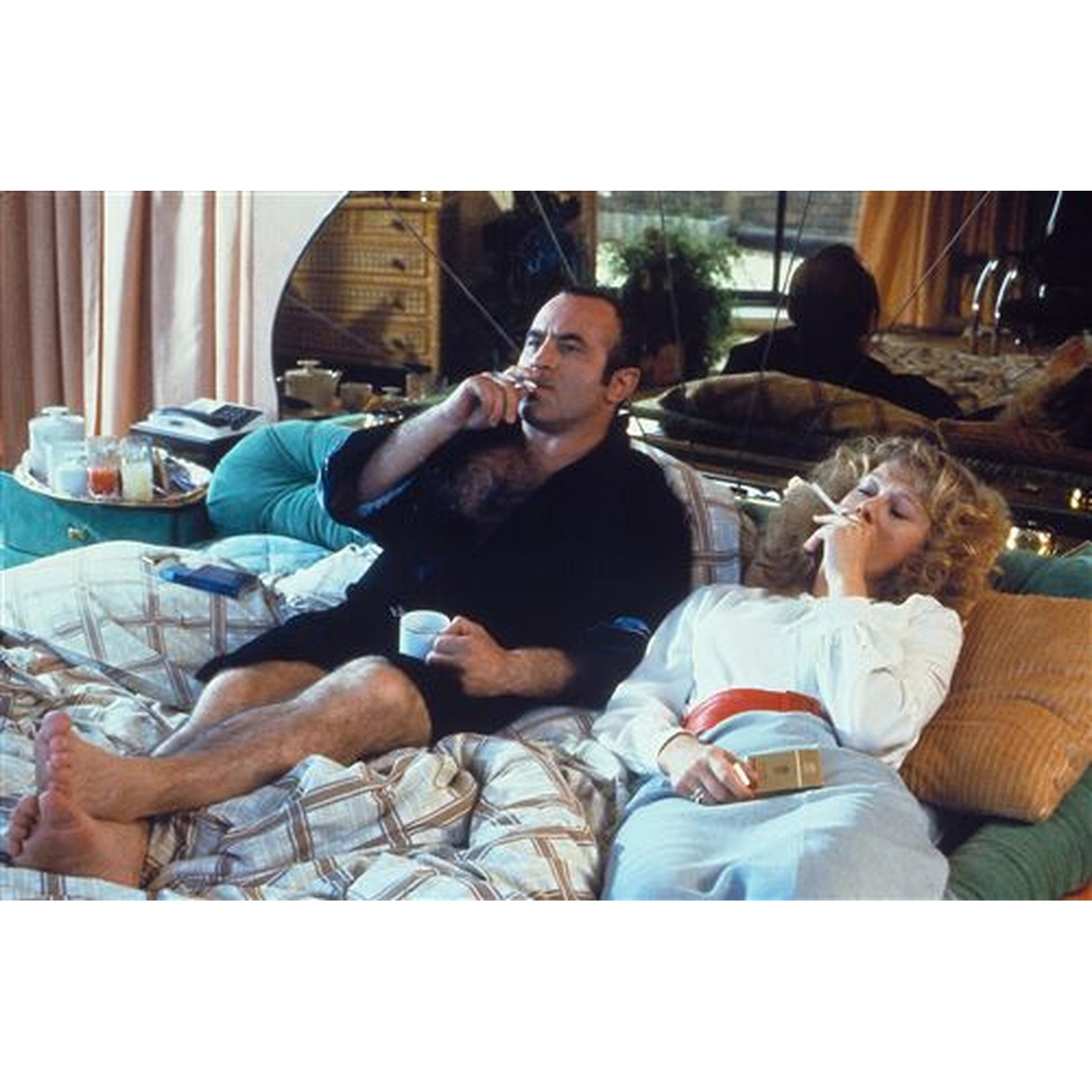 Un homme et une femme sont allongés sur un lit, fumant. L'homme tient une tasse. La femme porte une chemise blanche et une jupe bleue. Un plateau est à proximité.