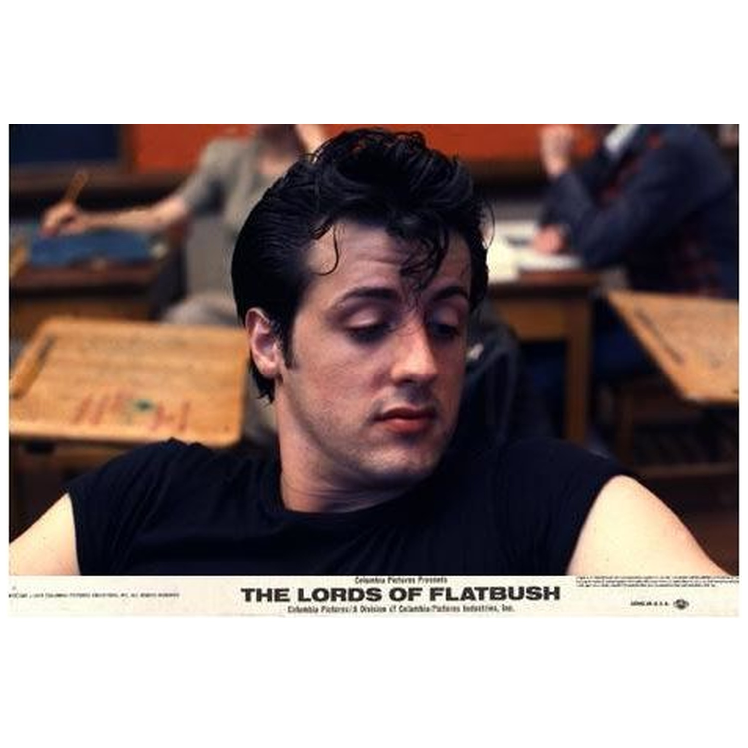 Sylvester Stallone dans The Lords of Flatbush. Il porte une chemise noire, regardant vers le bas. D'autres sont assis à des bureaux en arrière-plan.