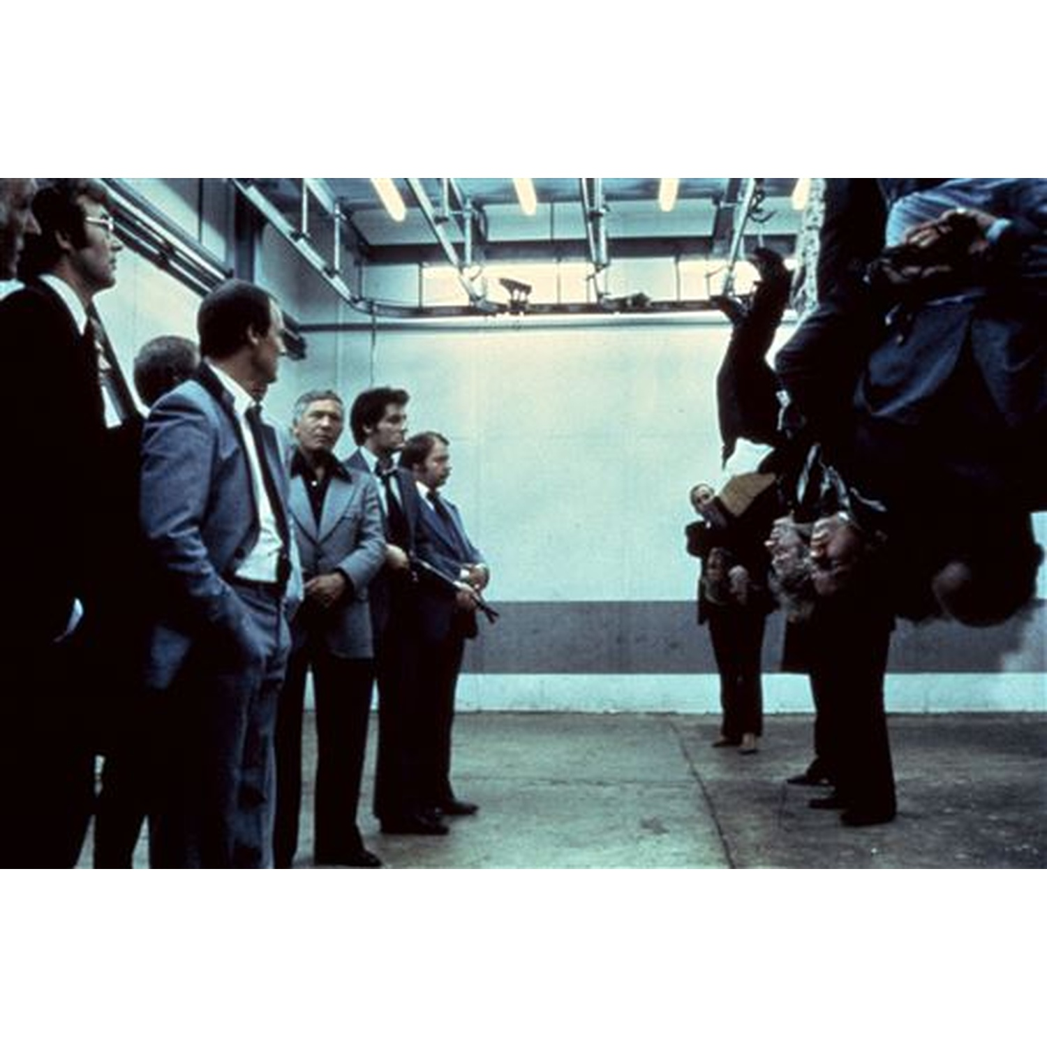 Men in suits stand in a room. One group looks at men hanging upside down.