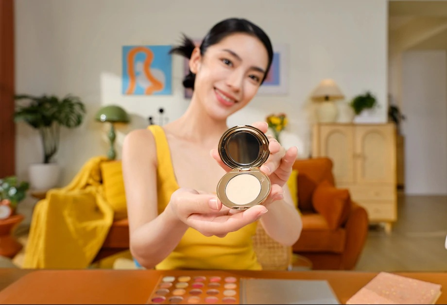 Kobieta w żółtym topie trzyma otwarty puder. Uśmiechnięta. Tło: sofa, sztuka, paleta do makijażu.