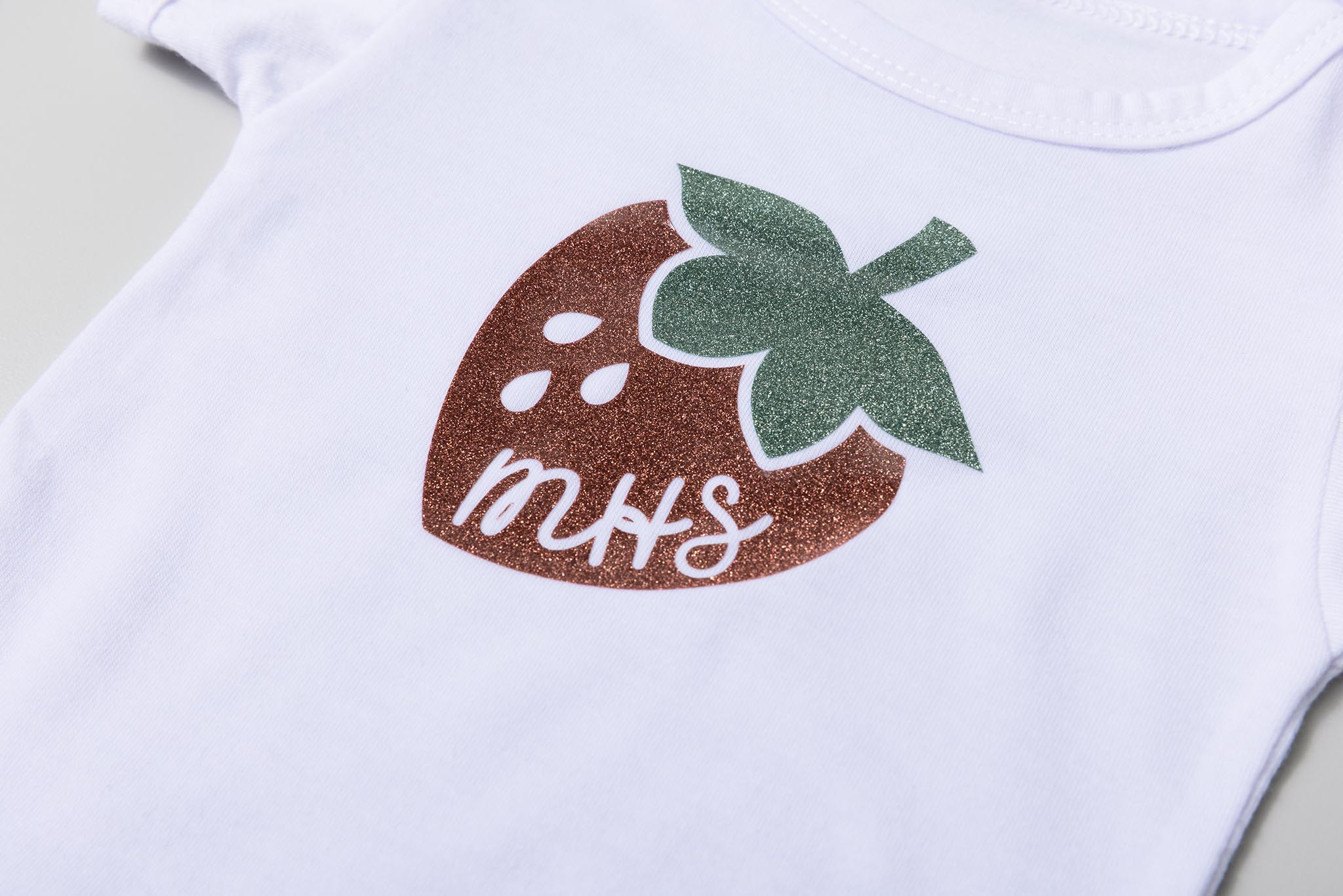 Weißer Baby-Body mit glitzerndem Erdbeer-Design. Die Erdbeere ist braun mit grünen Blättern, weiße Buchstaben sagen 'MHS'.