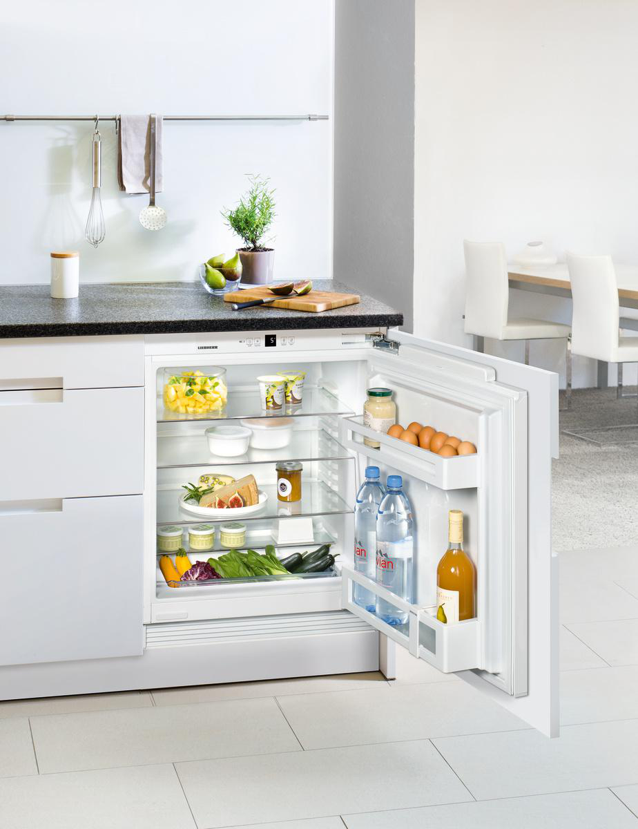 Offener Kühlschrank mit Obst, Gemüse und Getränken. Pflanze und Schneidebrett auf der Theke. Esstisch im Hintergrund sichtbar.