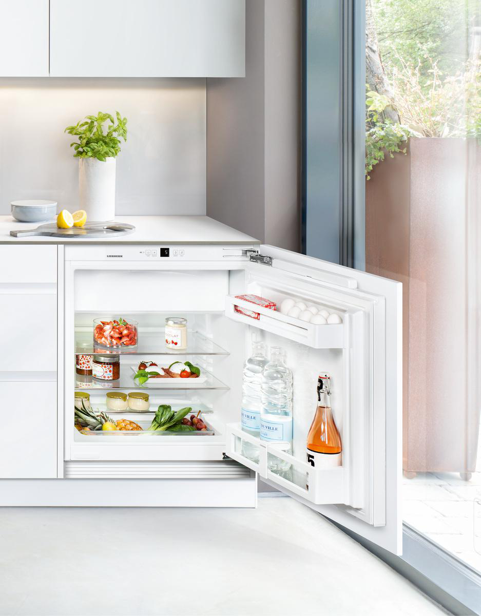 Offener weißer Kühlschrank mit Lebensmitteln und Getränken. Glastür zeigt Früchte, Gemüse, Wasserflaschen und eine Flasche Orangensaft.