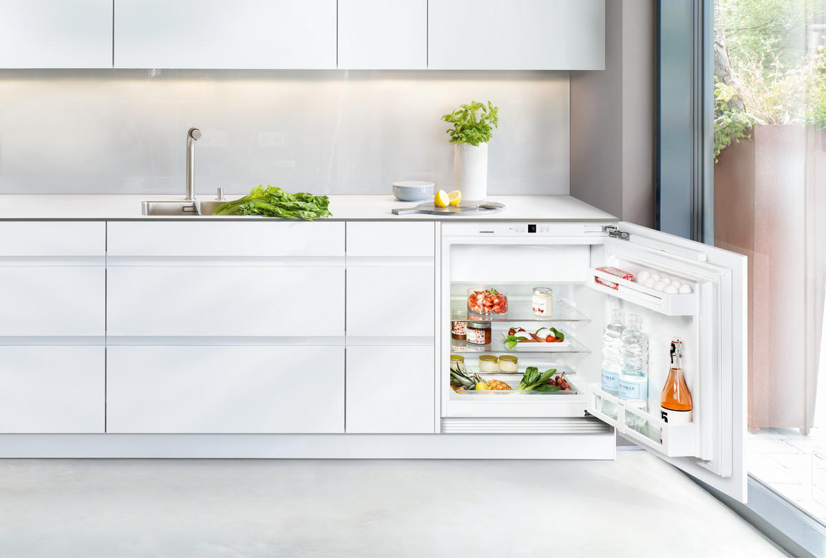 Eine moderne Küche mit einem teilweise geöffneten Kühlschrank voller Lebensmittel. Frisches Gemüse und ein Waschbecken sind sichtbar.