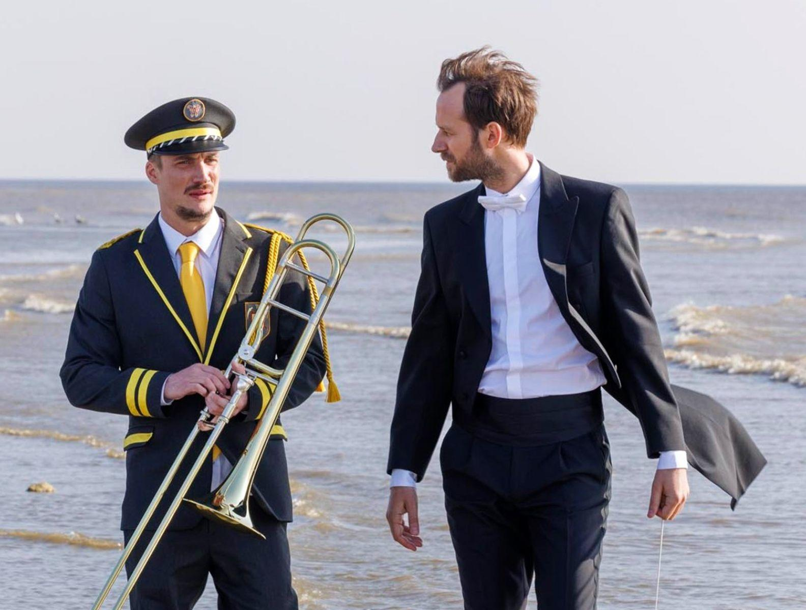 Zwei Männer am Strand. Einer in Uniform mit Posaune. Der andere im Anzug mit Fliege. Meer im Hintergrund.