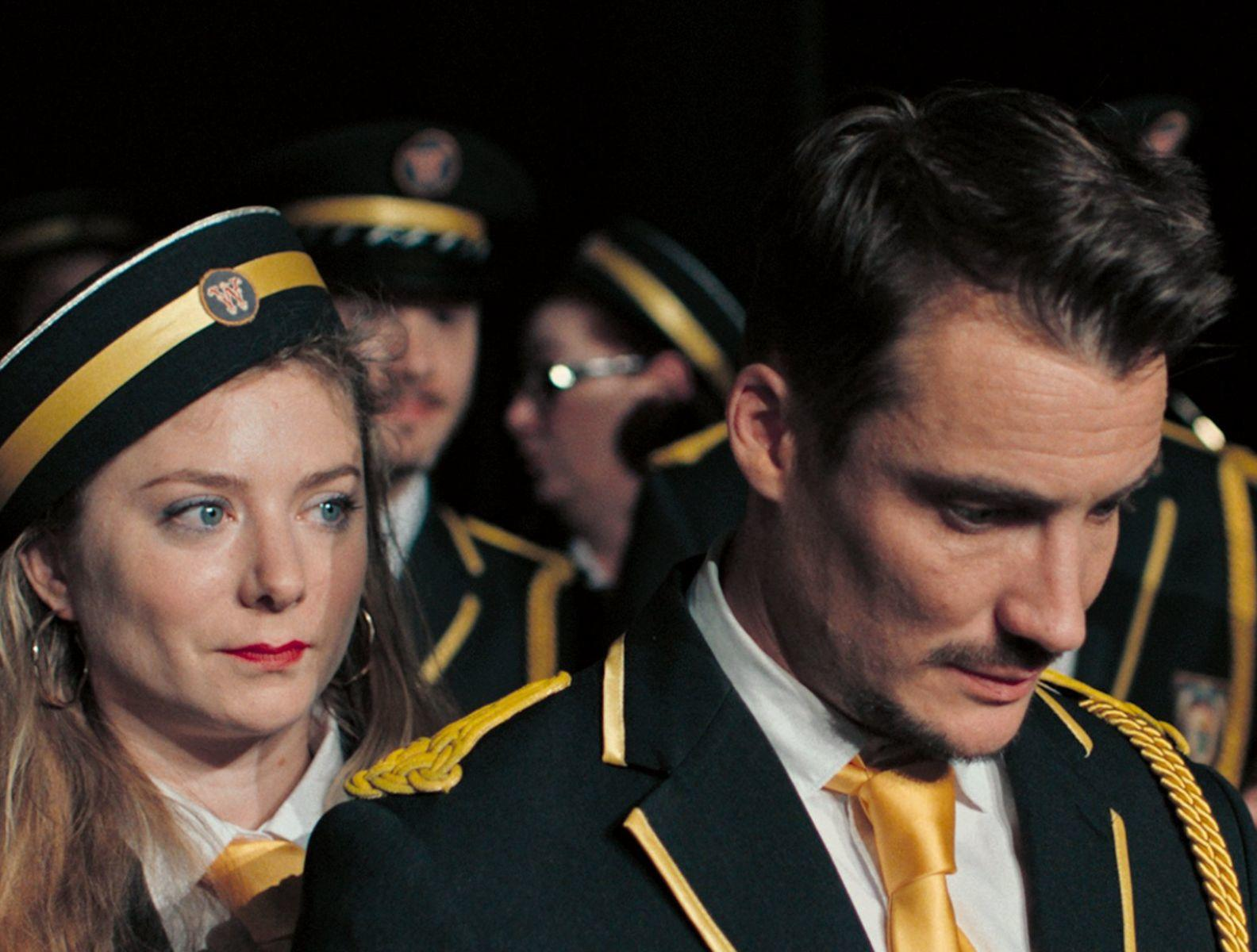 Zwei Personen in Uniform. Eine Frau mit Hut und ein Mann mit Schnurrbart. Andere sind im Hintergrund. Schwarze Uniformen mit goldenen Akzenten.