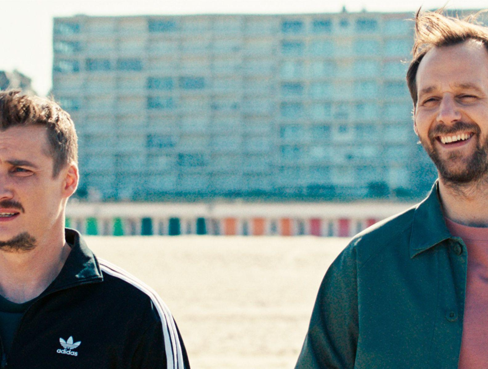 Zwei Männer am Strand lächeln. Einer trägt eine schwarze Adidas-Jacke, der andere ein grünes Hemd. Im Hintergrund ein Gebäude und bunte Strandhütten.