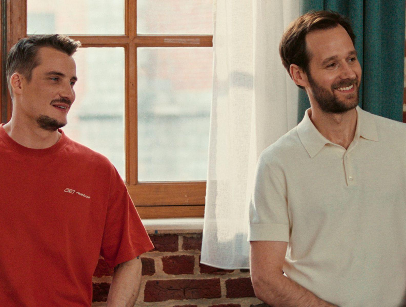 Zwei Männer stehen in der Nähe eines Fensters. Einer trägt ein rotes Hemd, der andere ein cremefarbenes Polohemd. Beide lächeln. Ziegelmauer und Vorhang sind sichtbar.