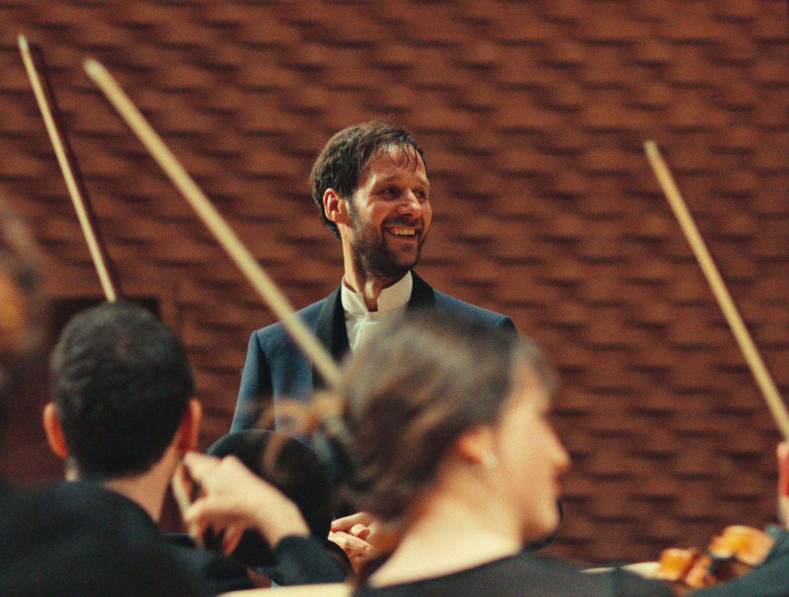 Ein Dirigent lächelt beim Leiten eines Orchesters. Bögen sind sichtbar. Ziegelmauer im Hintergrund.