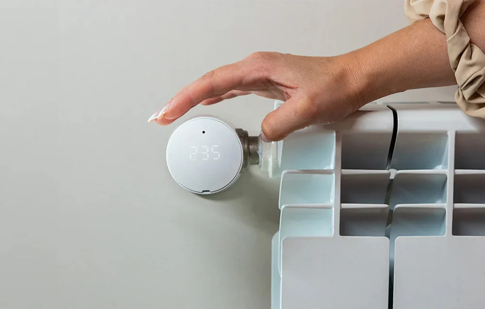 Ręka reguluje biały inteligentny termostat na grzejniku. Wyświetlacz pokazuje numer 235. Grzejnik jest biały.