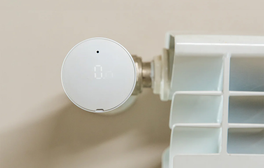 Biały inteligentny termostat z cyfrowym wyświetlaczem pokazującym '0'. Jest podłączony do białego grzejnika, na tle beżowej ściany.