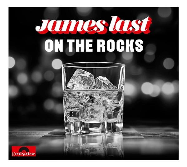 Ein Glas mit Eiswürfeln steht auf einer reflektierenden Oberfläche. Der Text lautet 'James Last ON THE ROCKS'. Hintergrund ist verschwommen.