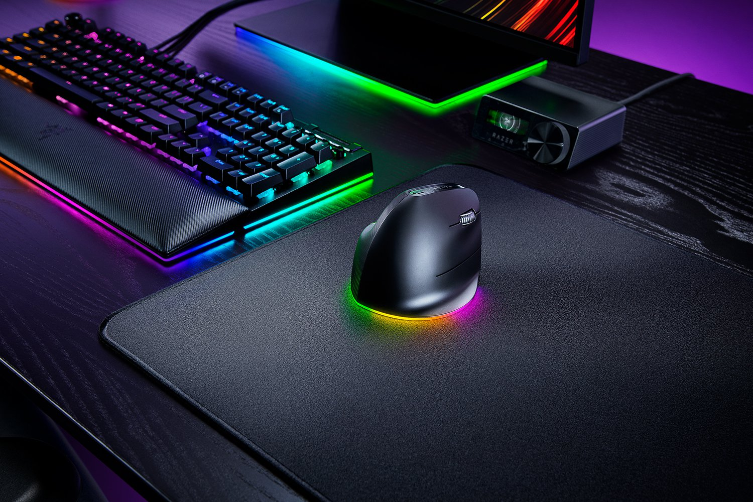 Un setup da gioco con tastiera, mouse verticale e monitor, tutti con illuminazione RGB colorata su una scrivania nera.