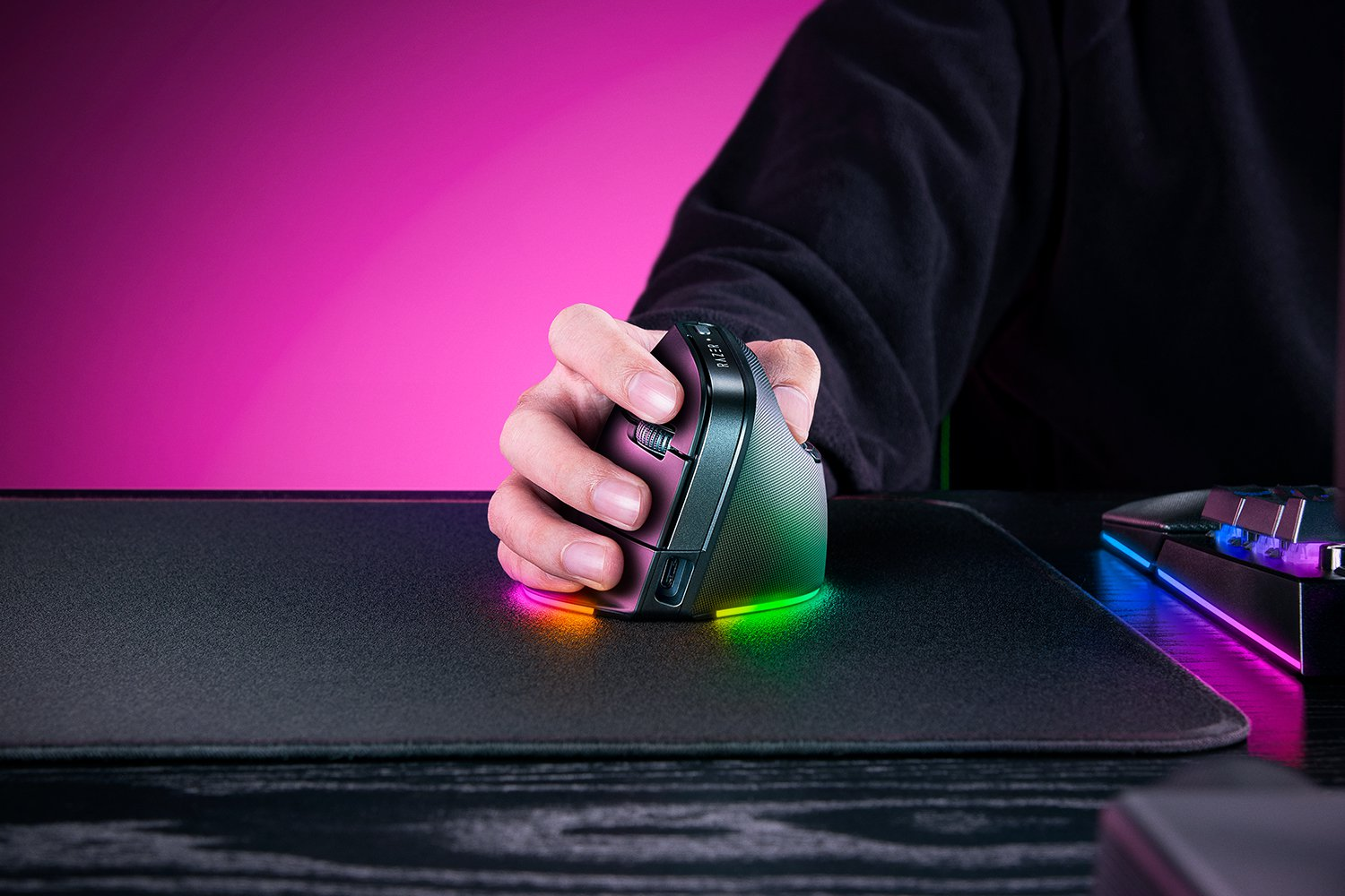 La mano di una persona è su un mouse viola e nero con luci colorate. Il mouse è su un tappetino nero.