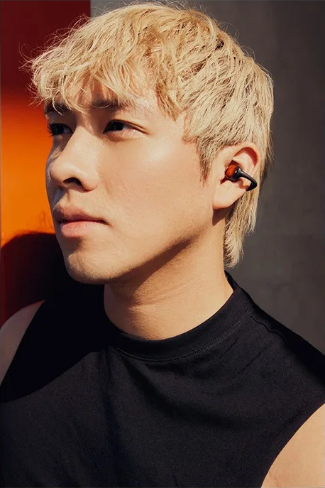 Un homme aux cheveux blonds portant des écouteurs et un débardeur noir. Le fond a des teintes orange et grises.
