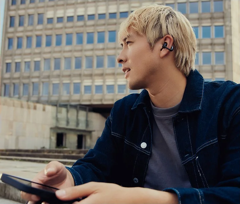 Ein Mann mit blonden Haaren, der Kopfhörer und eine Jeansjacke trägt, hält ein Telefon. Im Hintergrund ist ein Gebäude zu sehen.