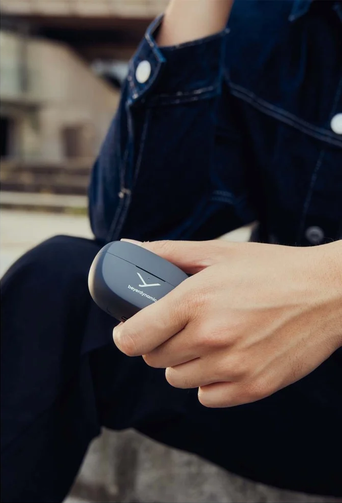 Eine Person hält eine graue Kopfhörerhülle mit dem Beyerdynamic-Logo. Sie trägt eine dunkle Jeansjacke.