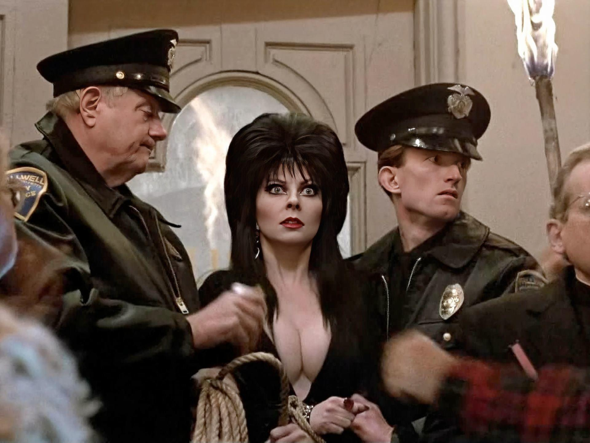 Elvira mit Polizisten. Sie trägt ein schwarzes Kleid. Zwei Offiziere in Uniform flankieren sie, ein anderer hält eine Fackel. Im Hintergrund ist ein Gebäude.