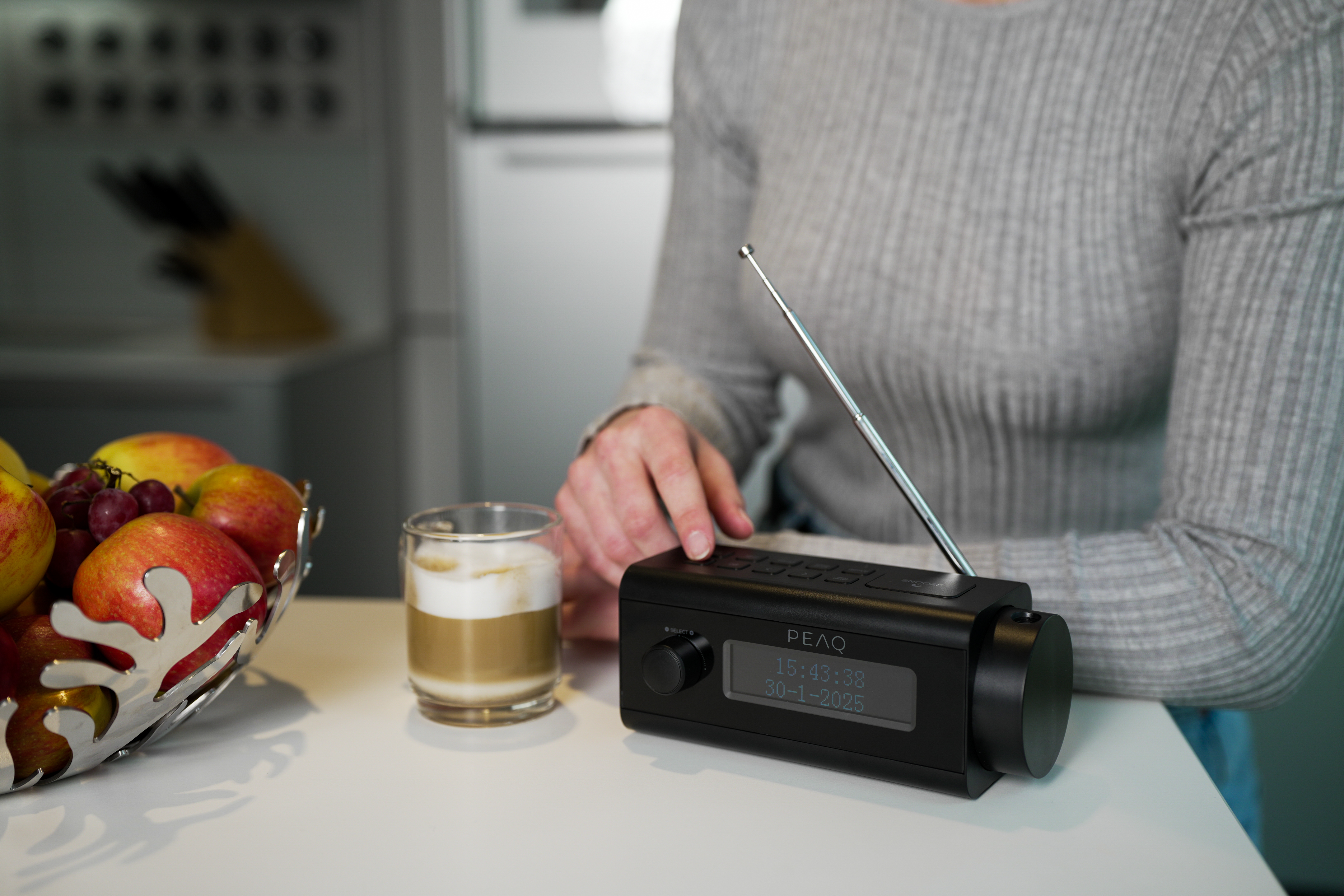 Une personne touche une radio noire avec affichage de l'heure. Des pommes et un café sont sur la table.