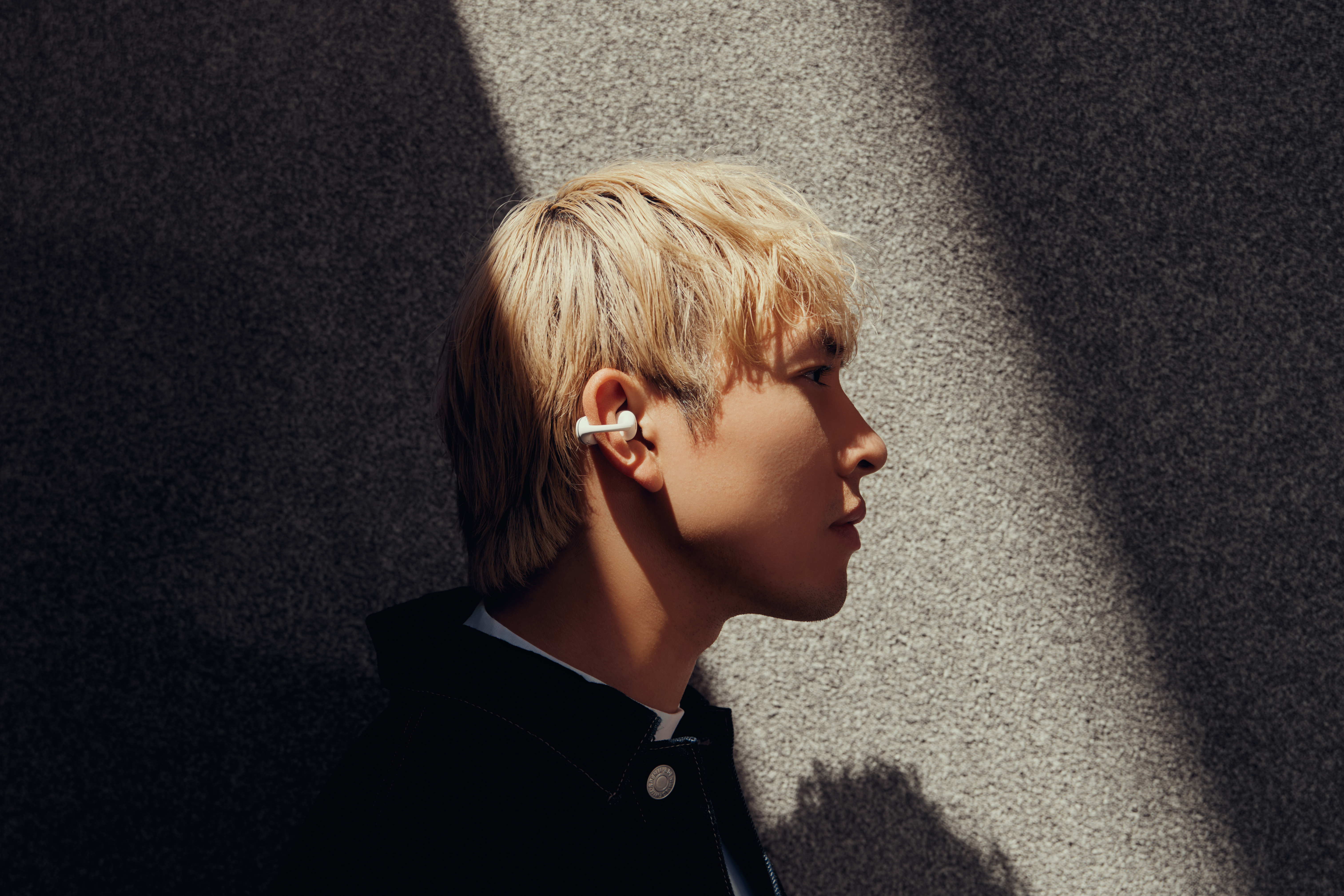 Ein Mann mit blonden Haaren, der weiße Ohrhörer trägt. Er ist im Profil vor einer strukturierten grauen Wand zu sehen, die von Sonnenlicht beleuchtet wird.