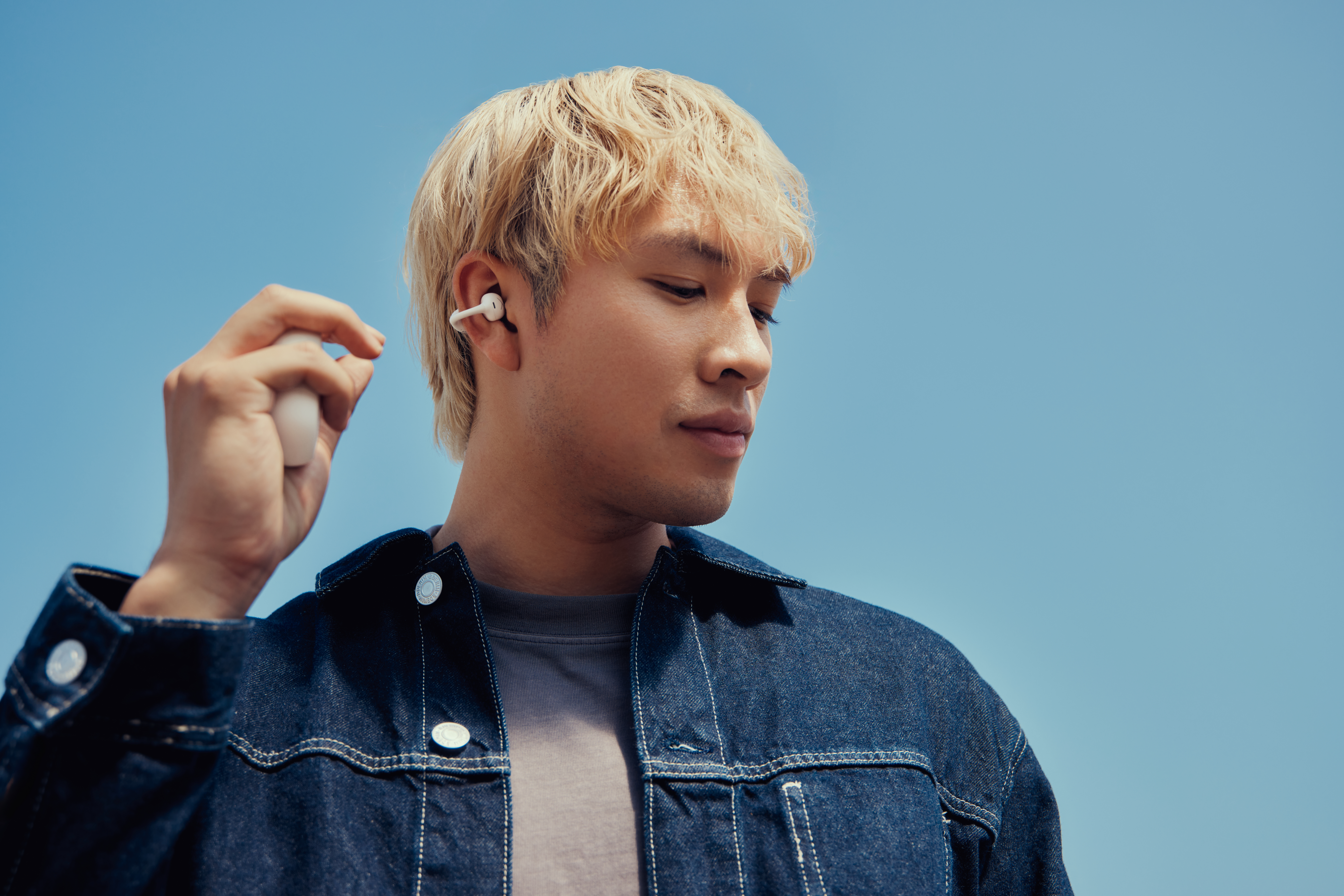 Ein Mann mit blonden Haaren setzt einen Ohrhörer ein. Er trägt eine Jeansjacke vor blauem Himmel.