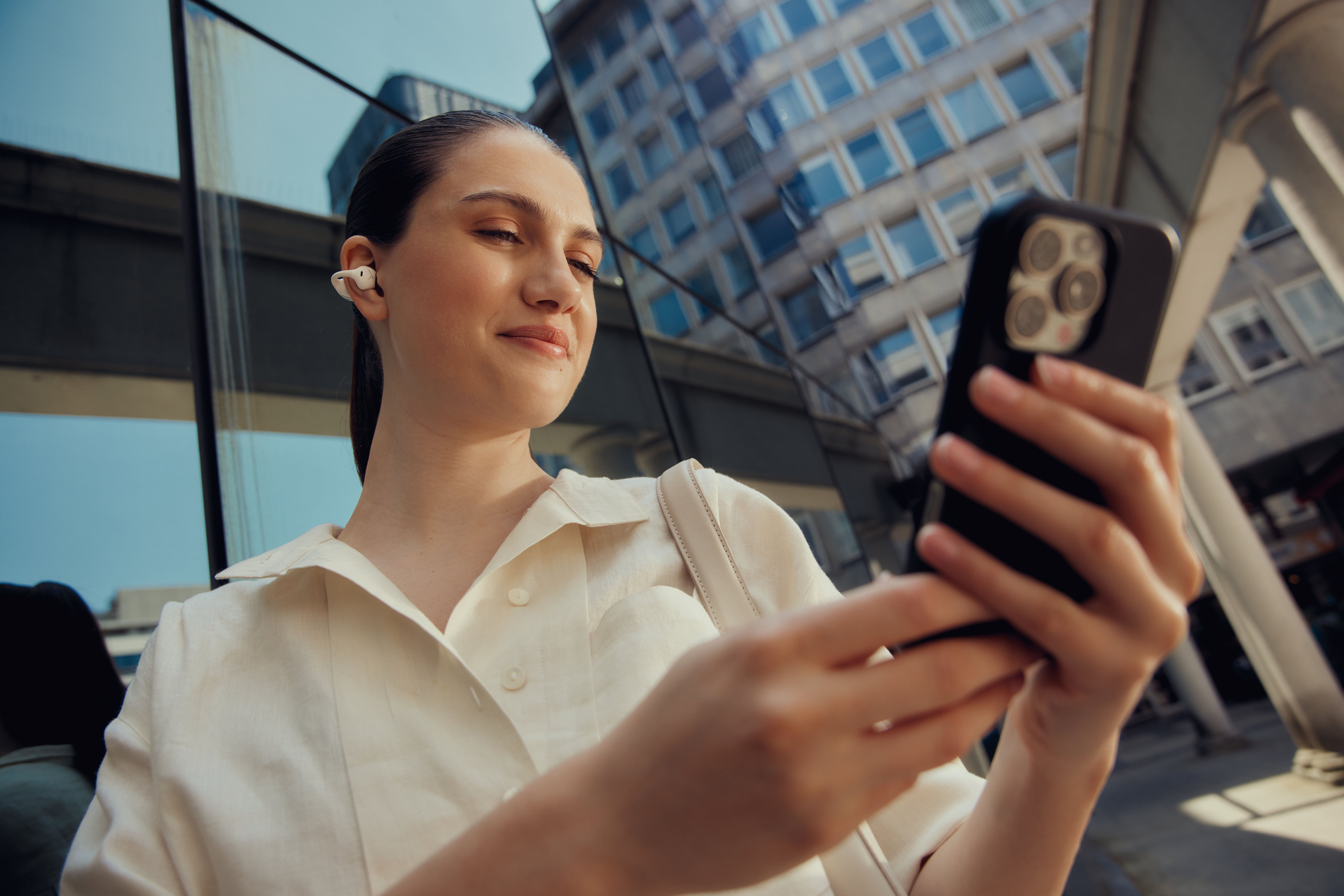 Frau mit Ohrhörern hält ein Telefon und schaut darauf. Gebäude im Hintergrund. Sonnig.