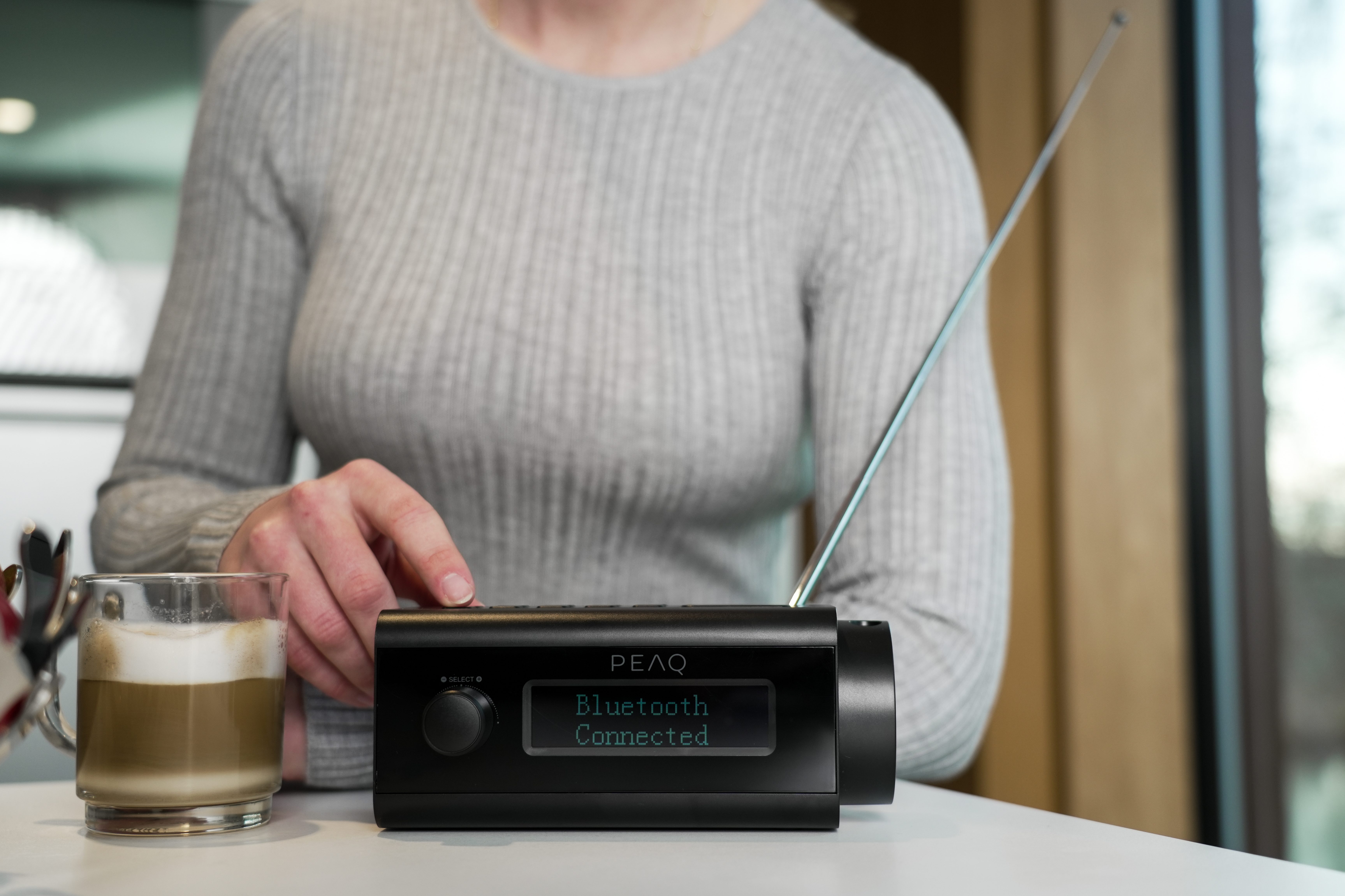 Une main appuie sur un bouton d'une radio noire. Une tasse de café est à gauche. L'écran affiche Bluetooth connecté.