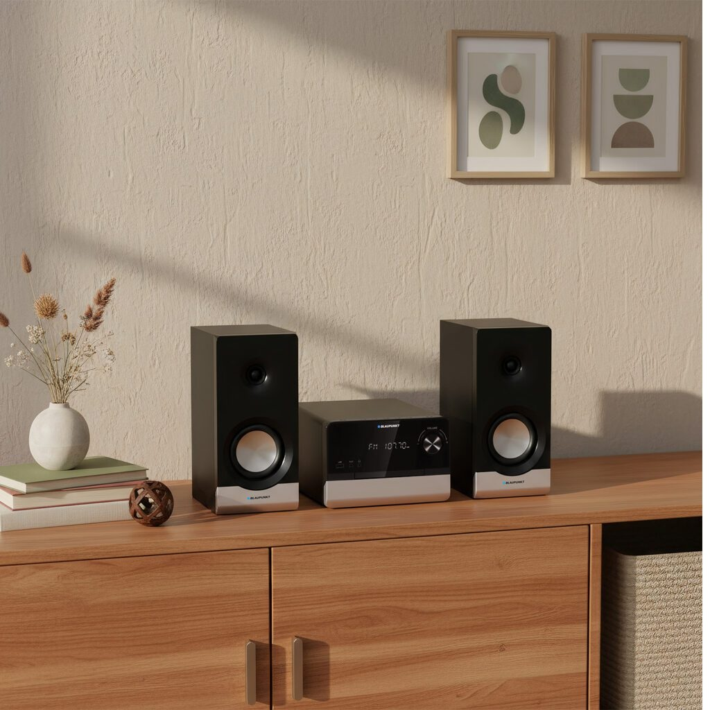 System stereo stoi na drewnianej szafce. Dwa czarne głośniki flankują jednostkę centralną. Na beżowej ścianie wiszą dzieła sztuki.