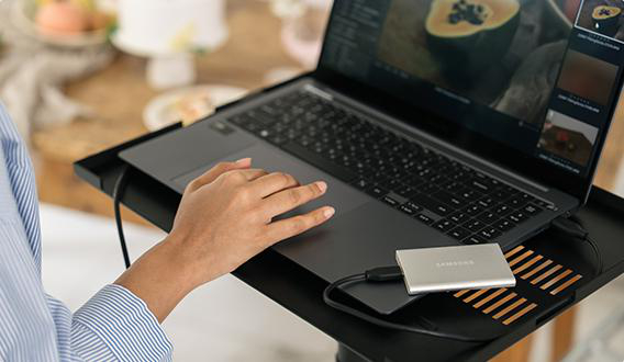 Eine Person benutzt einen Laptop auf einem schwarzen Ständer. Eine silberne Samsung Festplatte ist angeschlossen. Der Bildschirm zeigt eine Videoschnittstelle.
