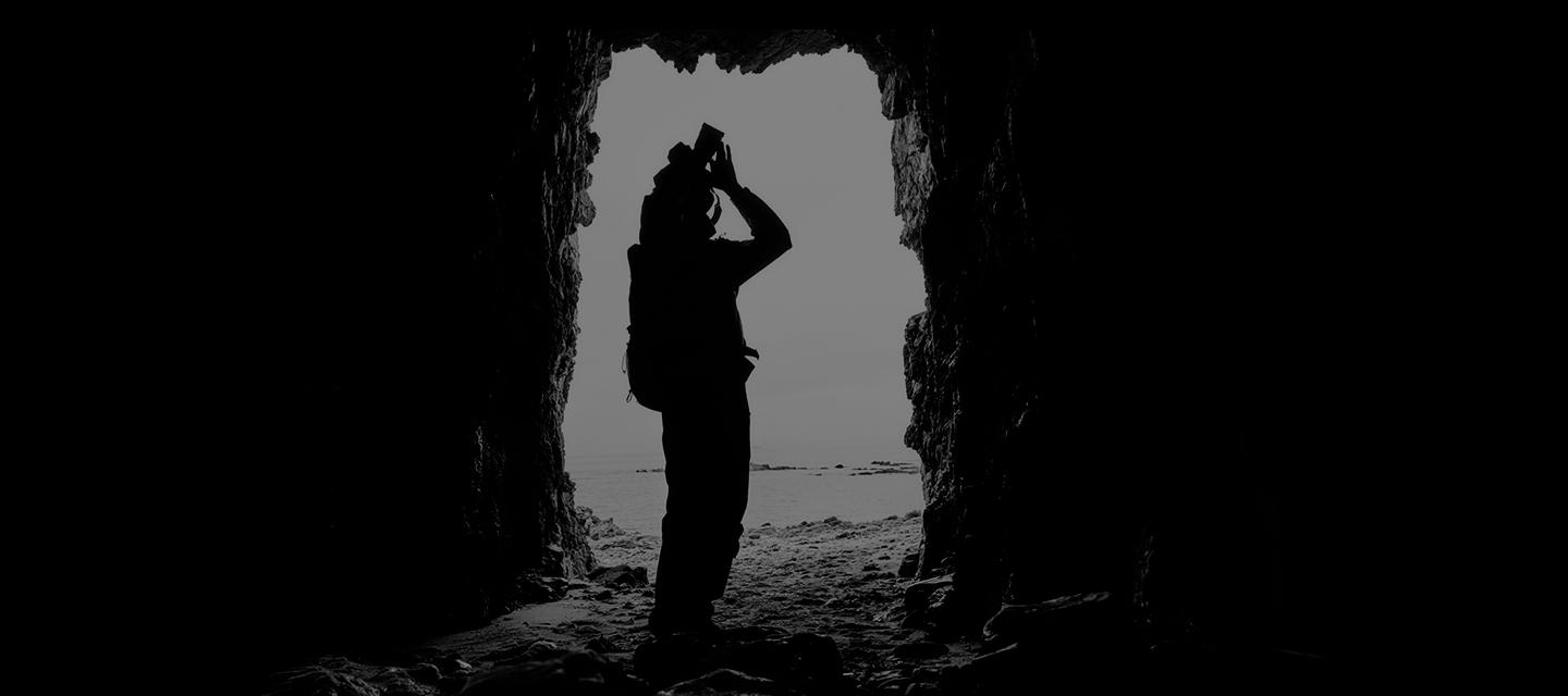 Silhouette einer Person in einer Höhlenöffnung. Sie hält einen Gegenstand hoch, möglicherweise eine Kamera, mit Blick nach draußen.